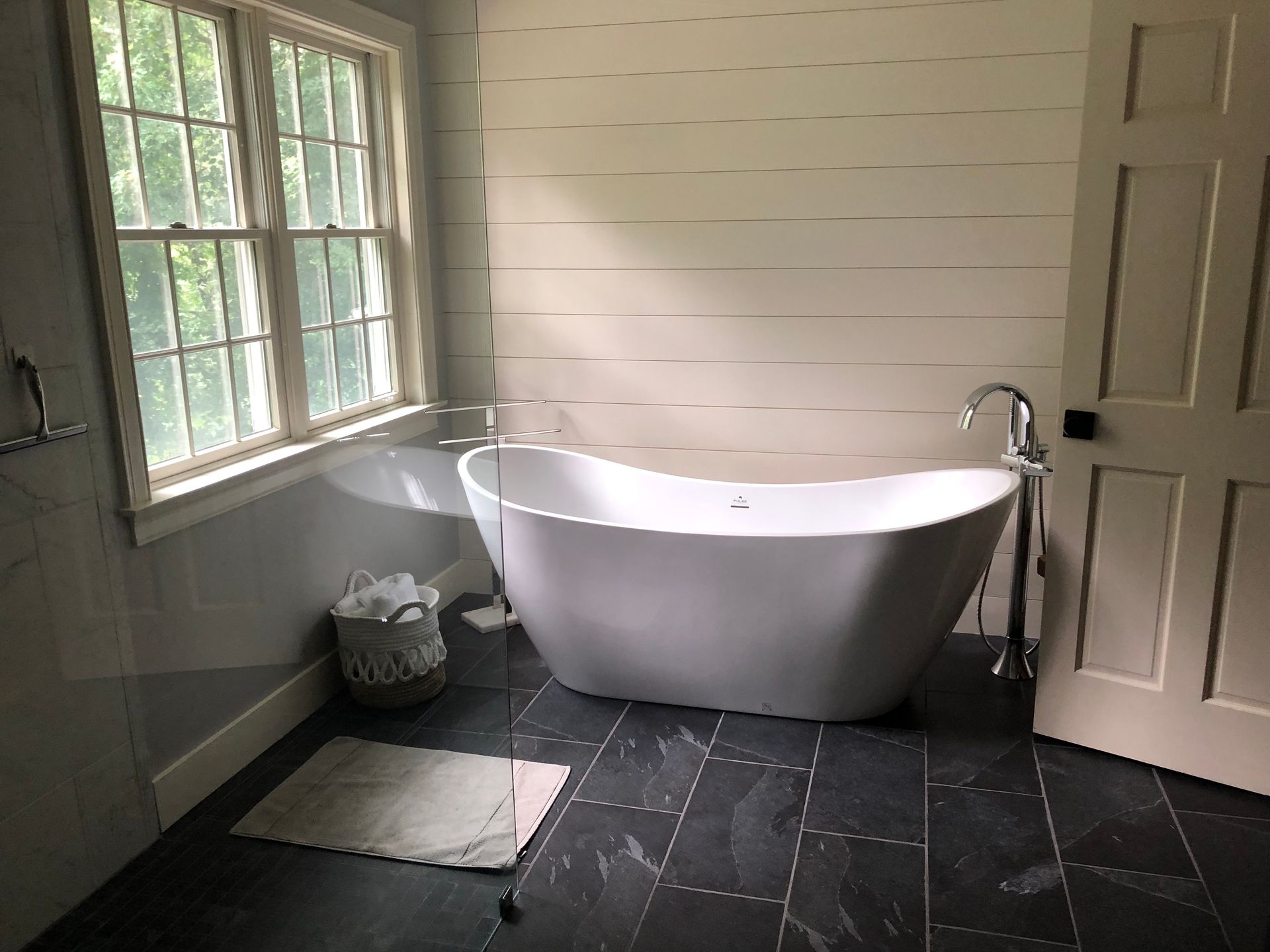 Bathroom with a white freestanding tub, window, dark tile floor, and shiplap wall.