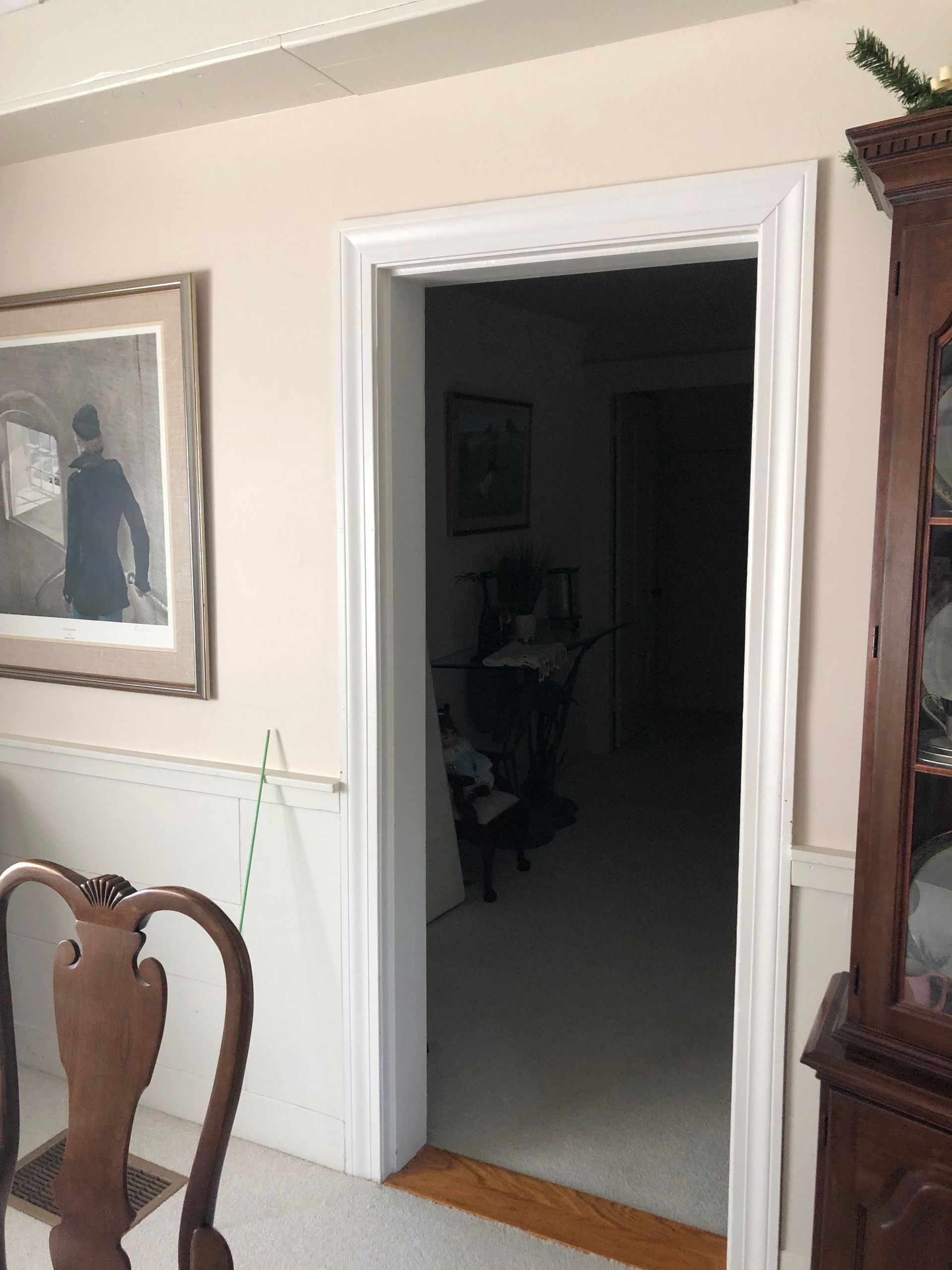 Doorway with white trim, leading to a dark hallway. Tan walls, painting, and wooden furniture on either side.