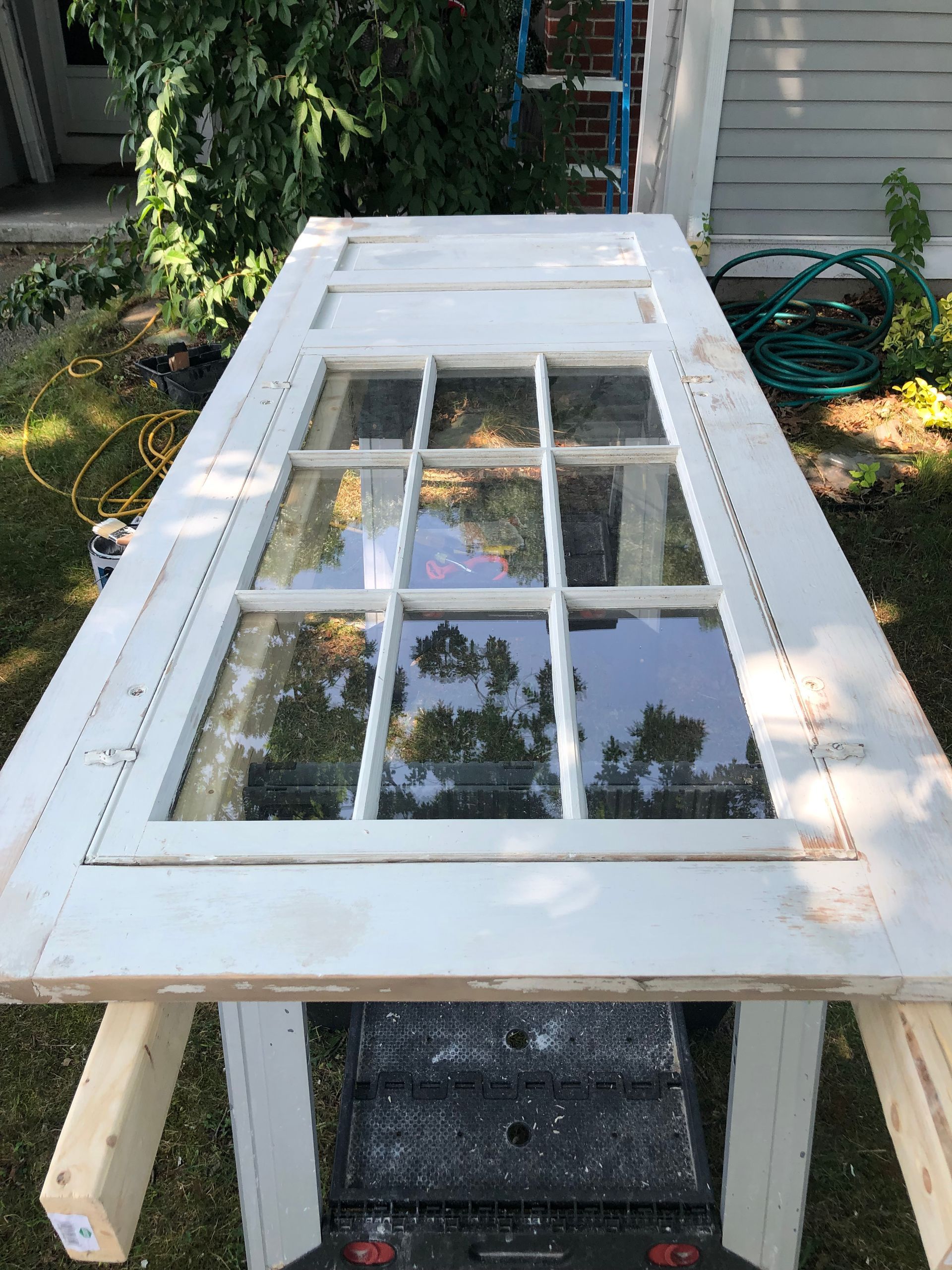 White door with glass panes resting on a worktable outdoors, with tools and a green hose nearby.