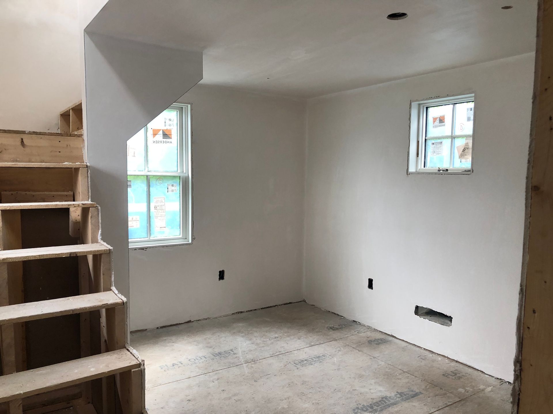 Interior room with unfinished concrete floor, two windows, newly plastered walls, and a wooden staircase.