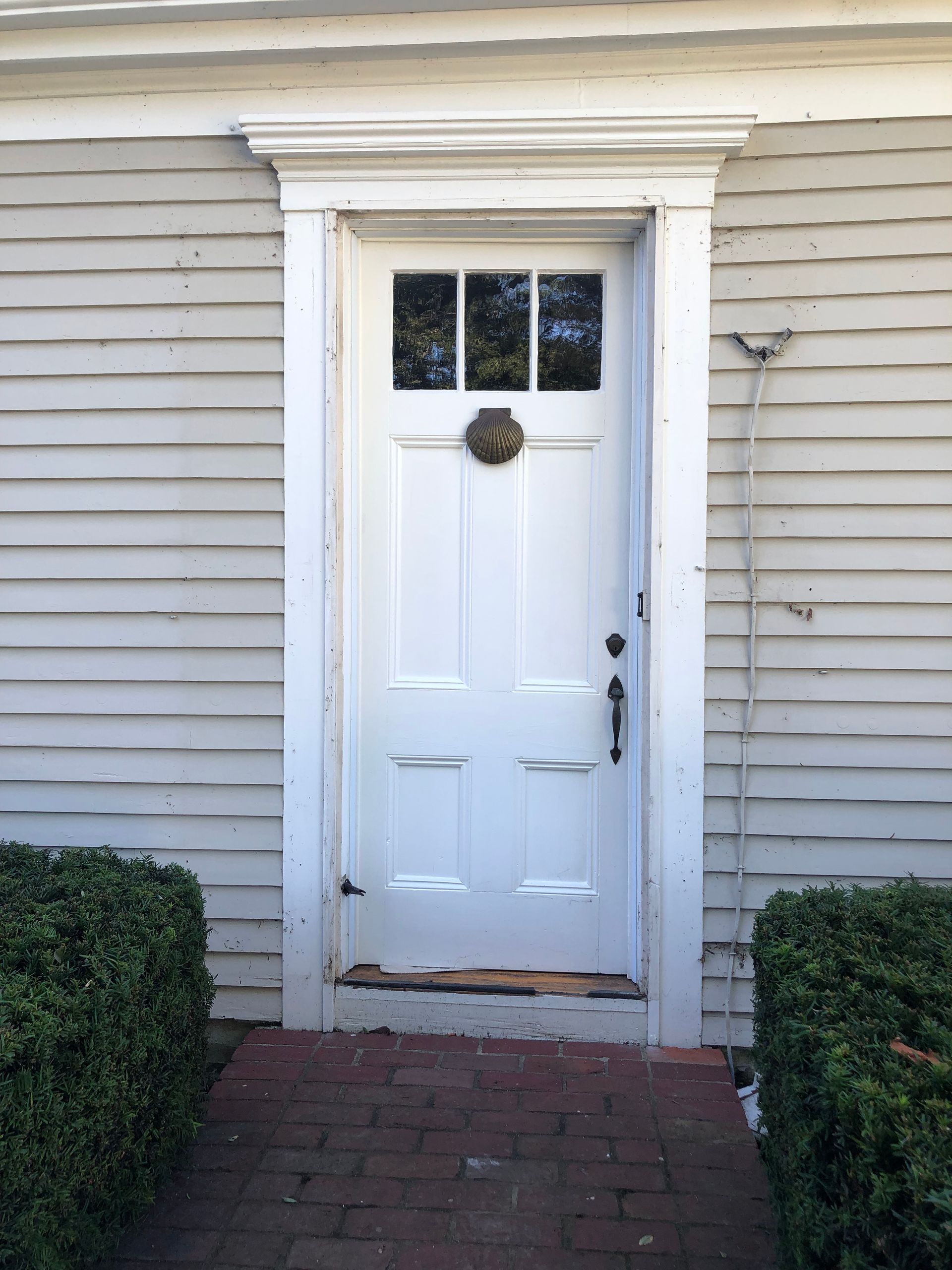 White door with small glass panes and knocker, framed by white trim, set in a gray clapboard wall.