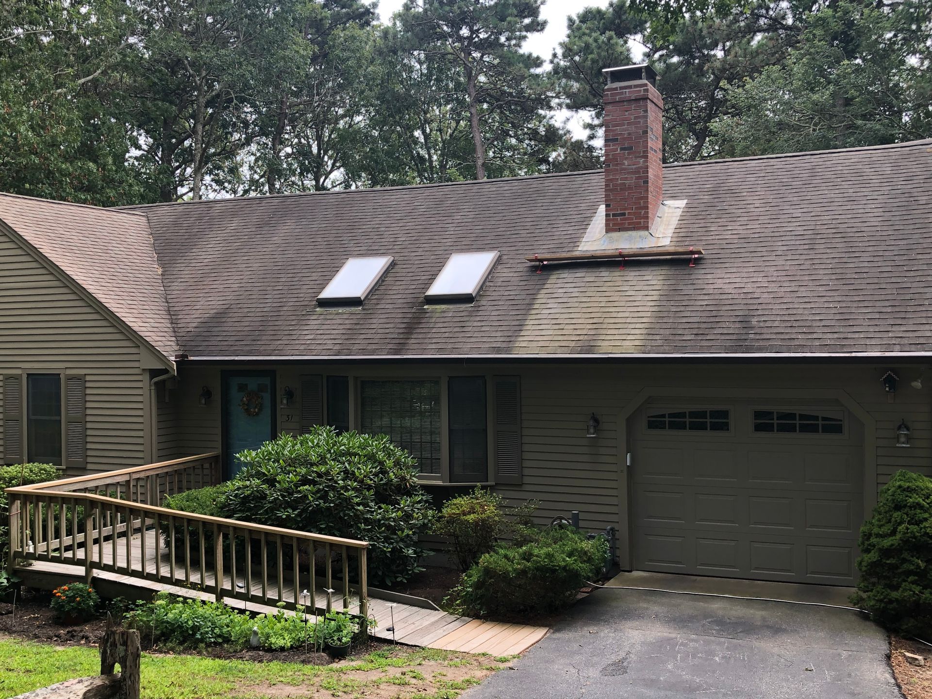 A single-story house with a wooden ramp and driveway, brown roof with swirl pattern, two skylights, and a brick chimney.