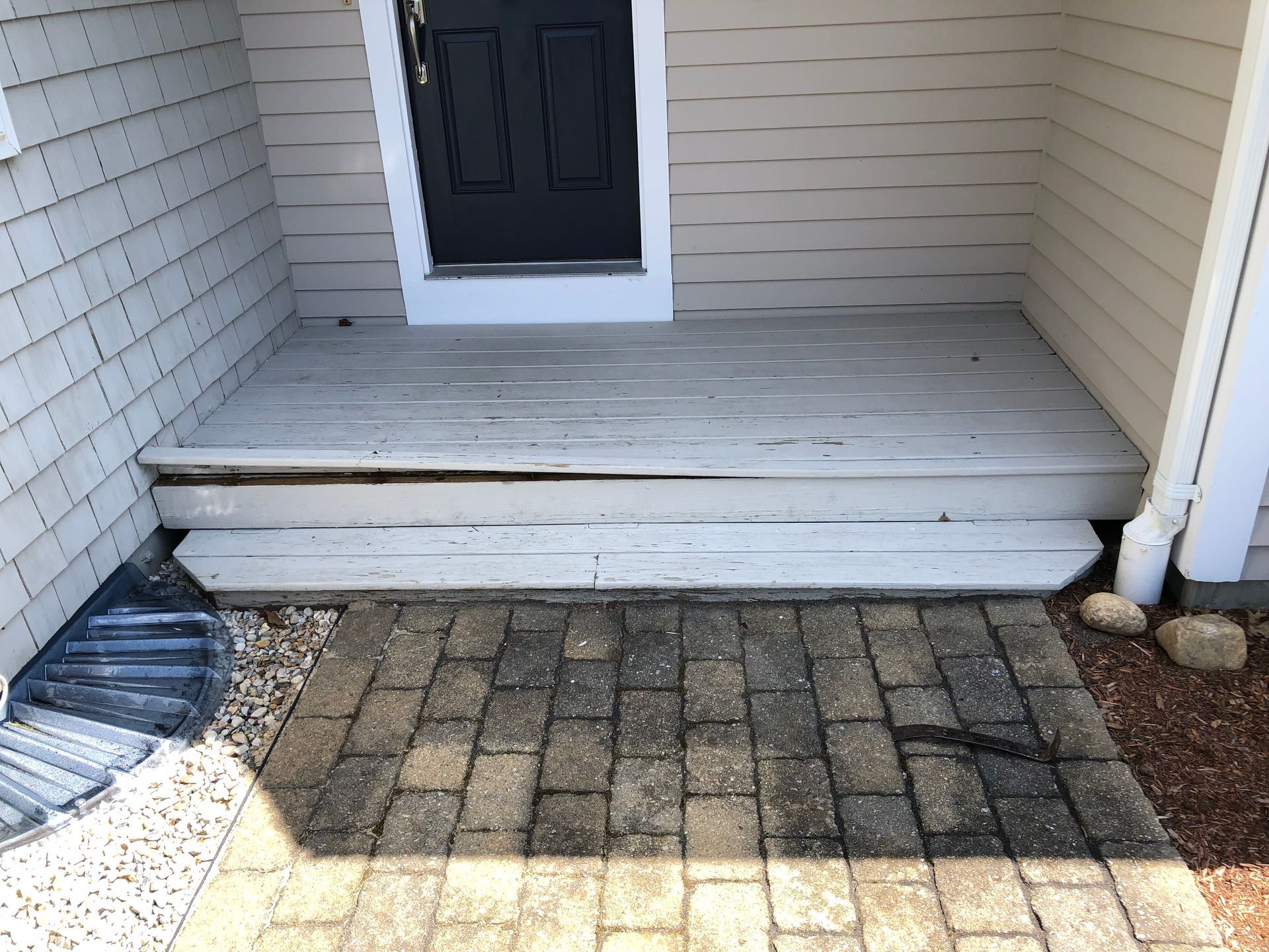 A weathered, white wooden porch with two steps leading to a black door, in front of a brick pathway.