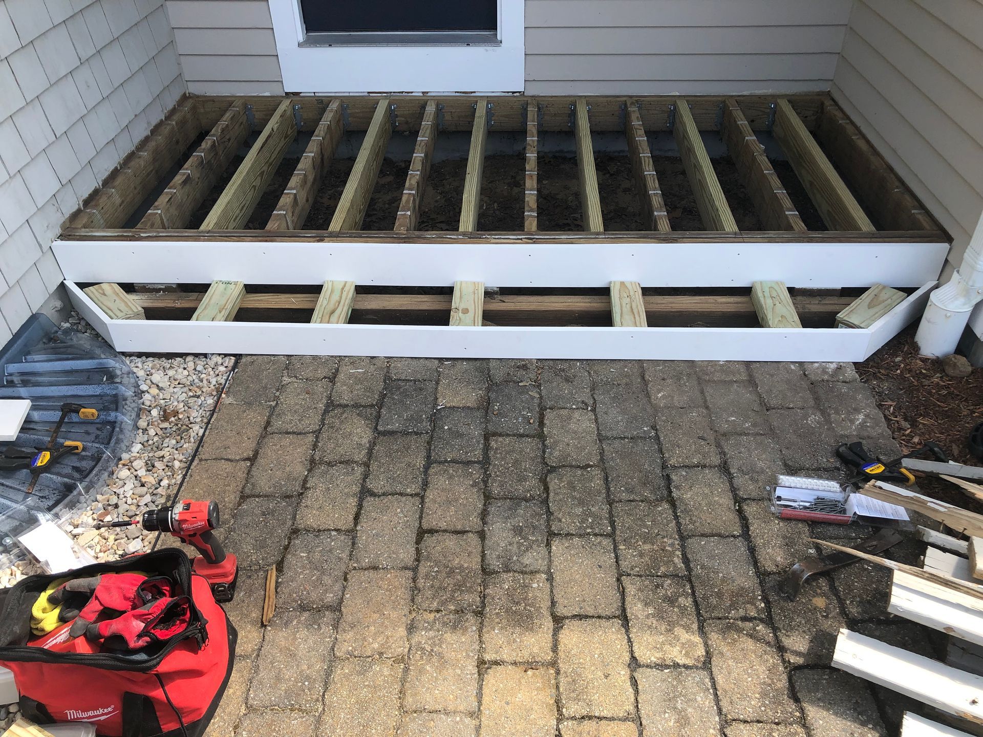 Framed deck structure under construction, white trim, open wooden frame on brick patio.