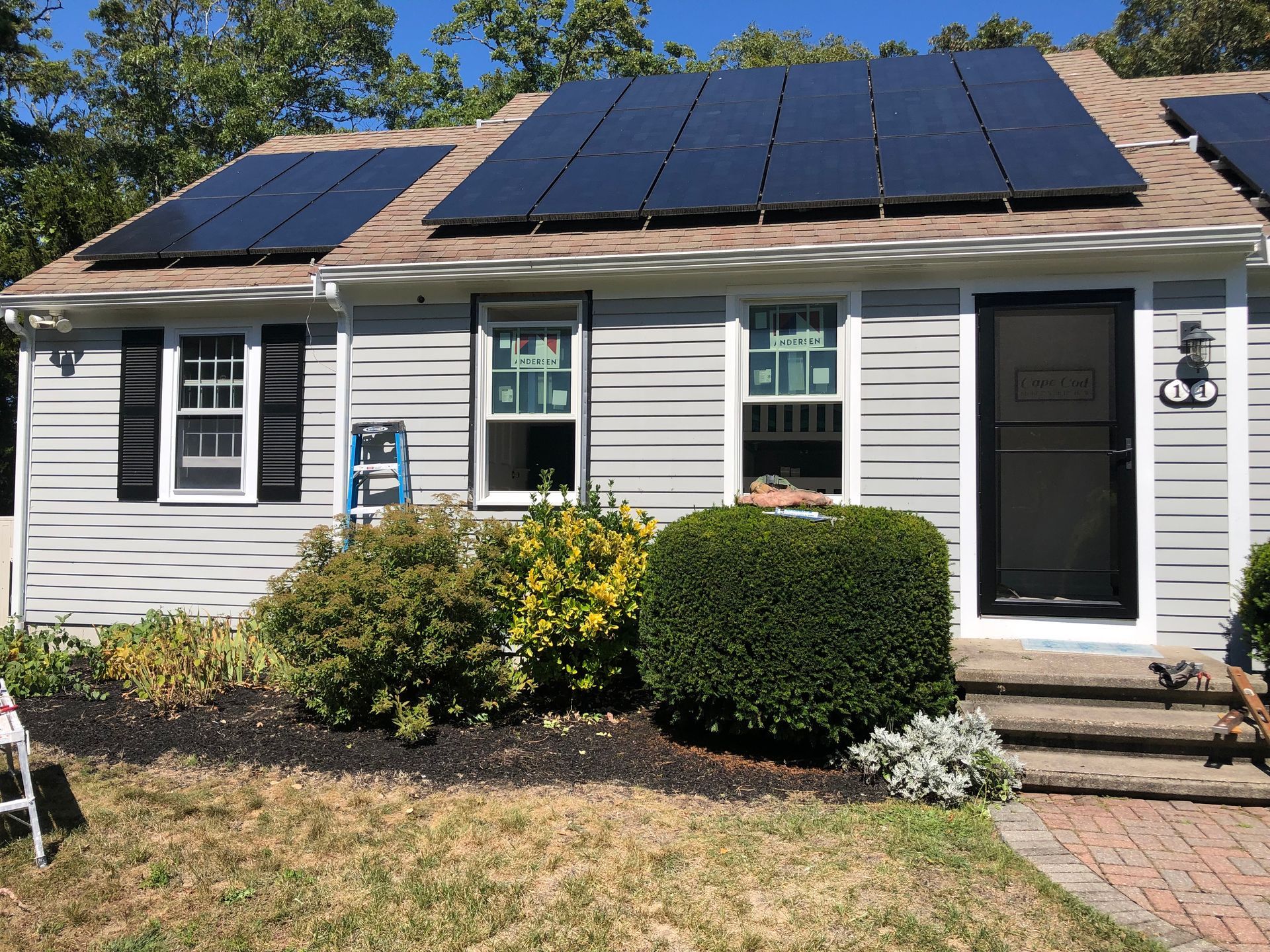 A gray house with solar panels on the roof. Front door and windows. Landscaping in front of the house.