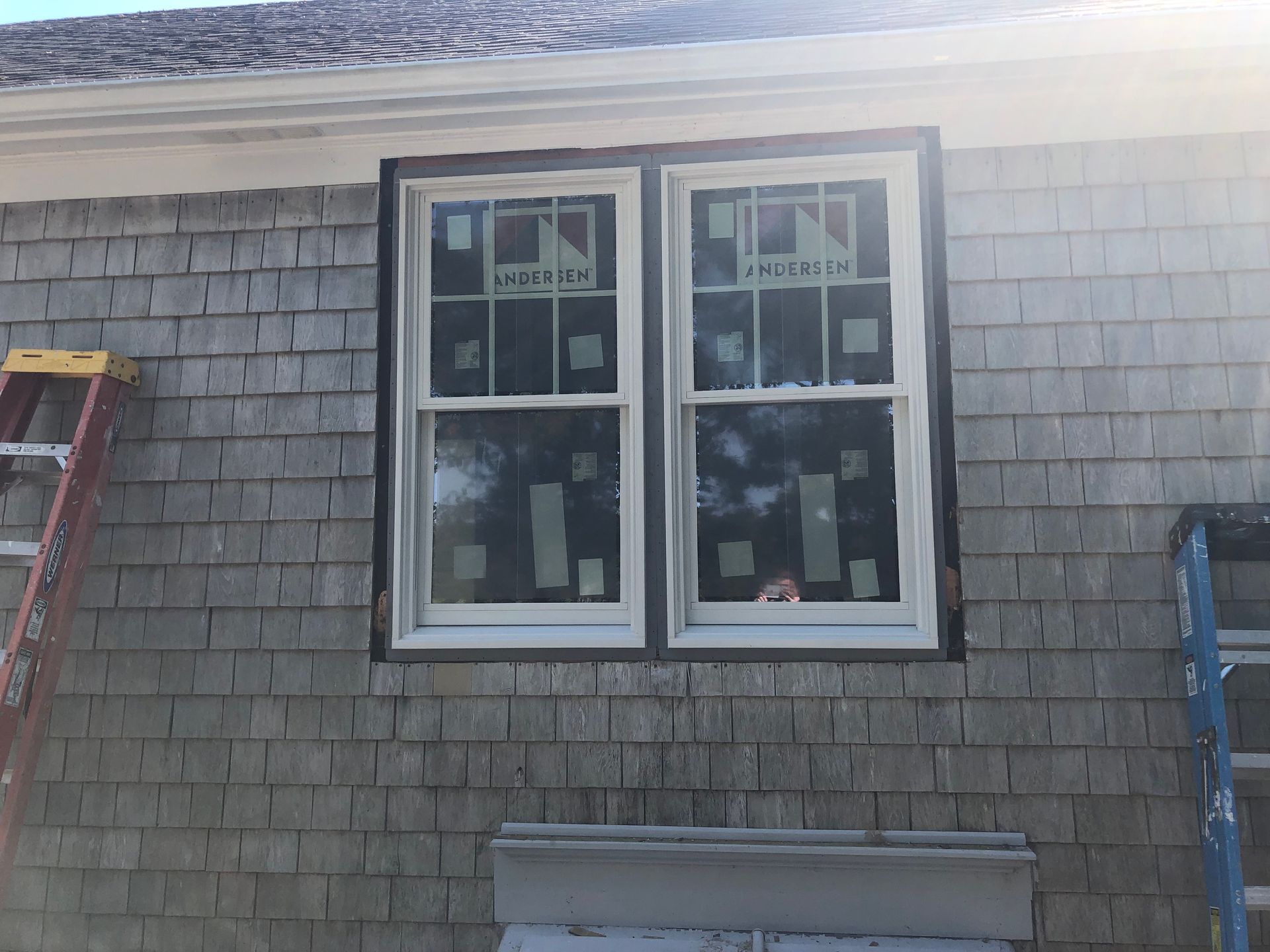 Two white double-hung windows installed in a gray shingled wall with a black trim; a ladder is on the left.