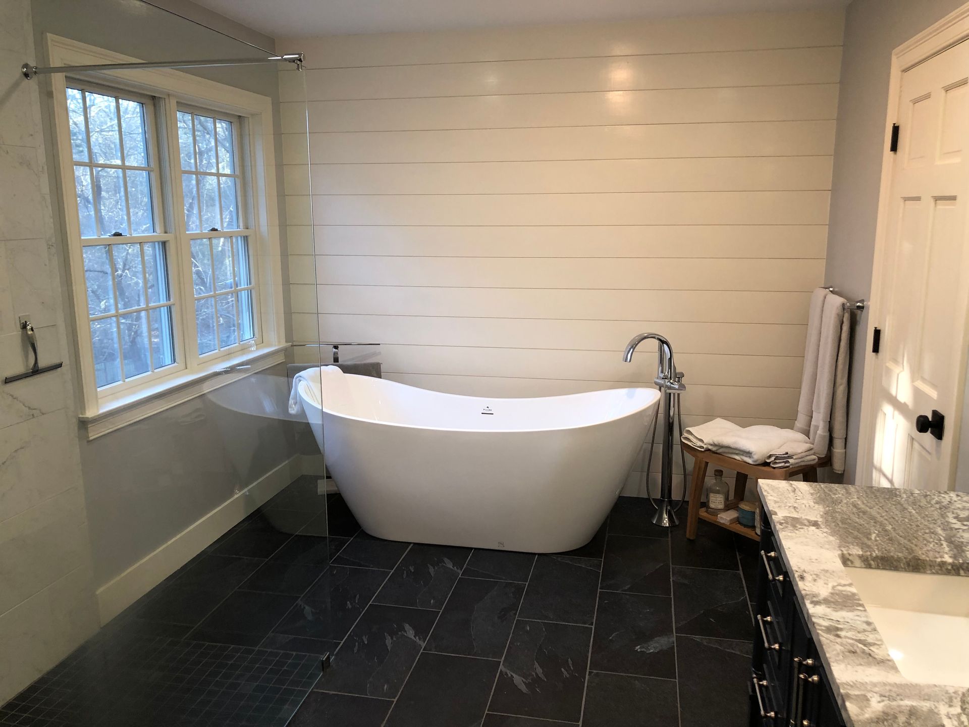 Bathroom with a white freestanding tub, wood-plank wall, dark tiled floor, and a window.