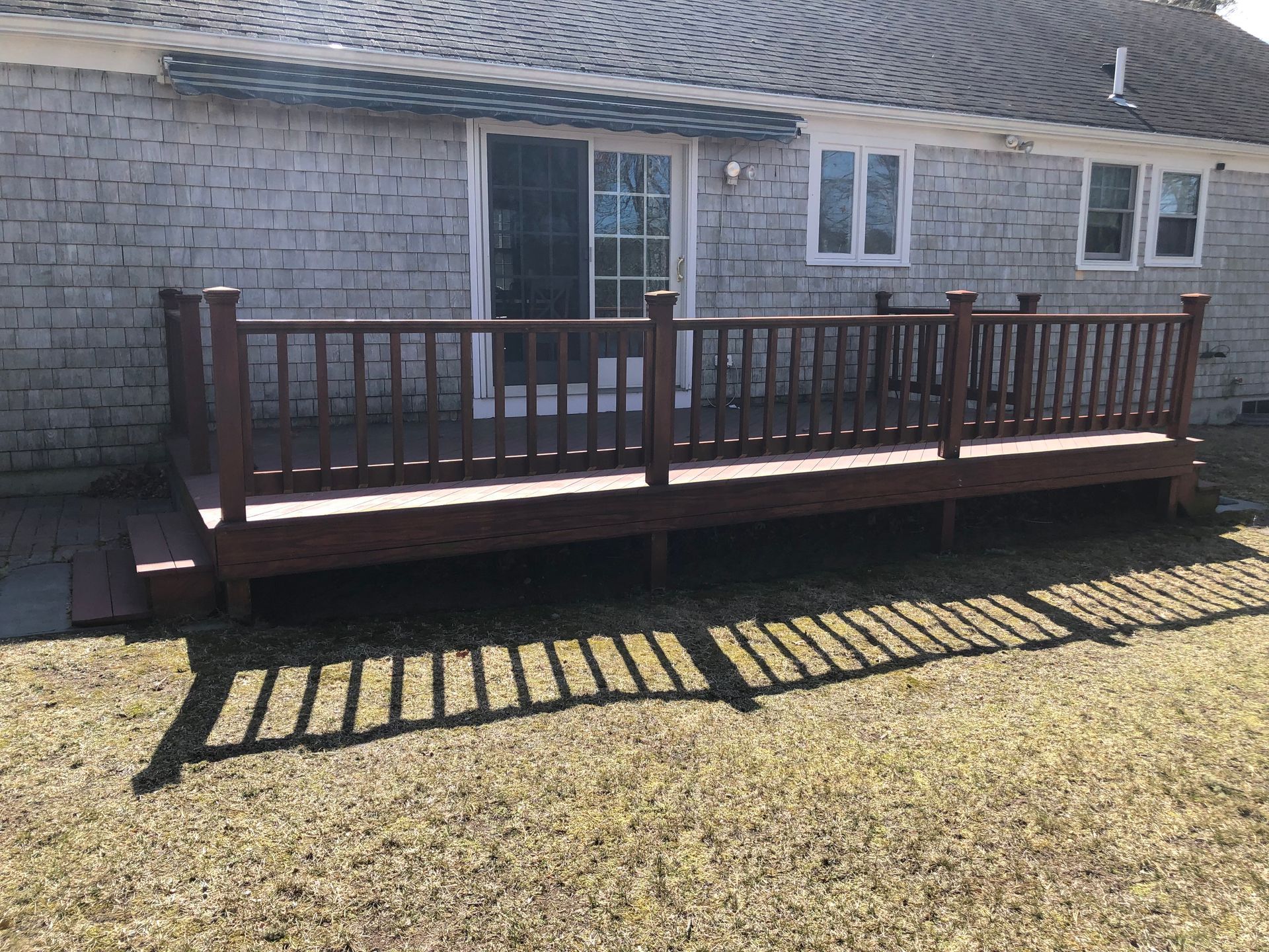 Wooden deck attached to a gray-sided house with a sliding door and windows. Brown railings and a ramp.