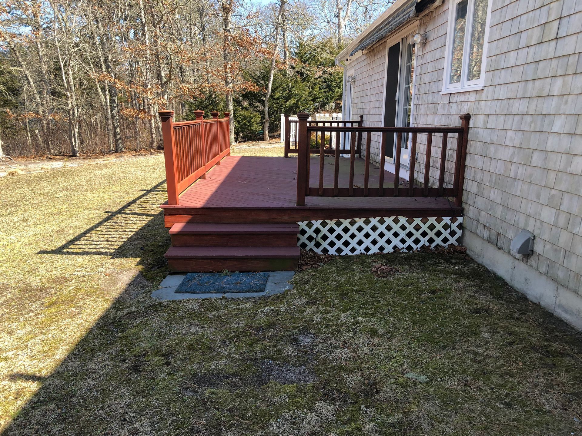 Red deck with steps attached to a light-colored house; white lattice, dead grass, and trees in the background.