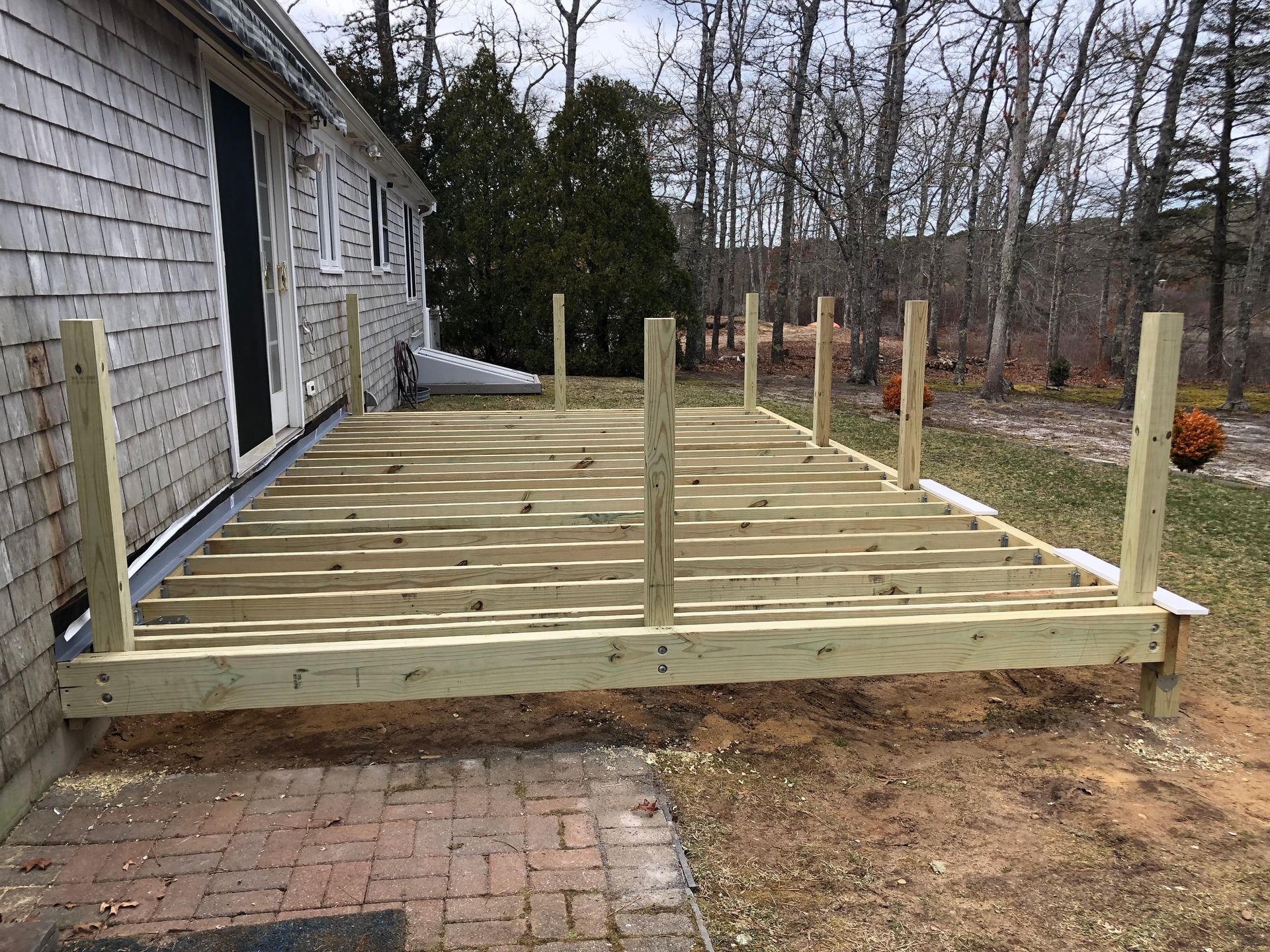 Wooden deck frame attached to a house with vertical posts for a railing, on a brick patio.