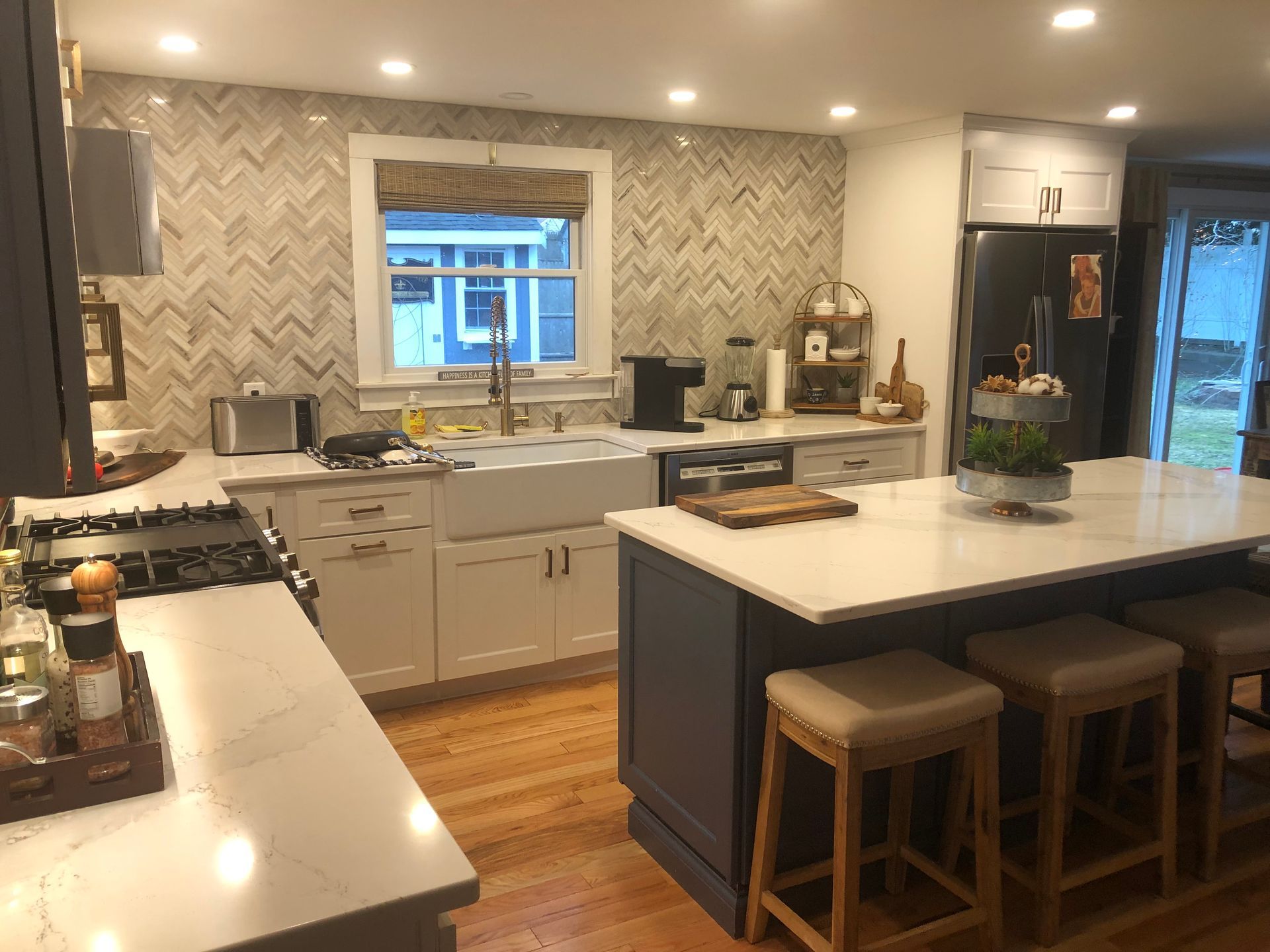 Kitchen with white cabinets, dark blue island, white countertops, herringbone backsplash, and wood floors.