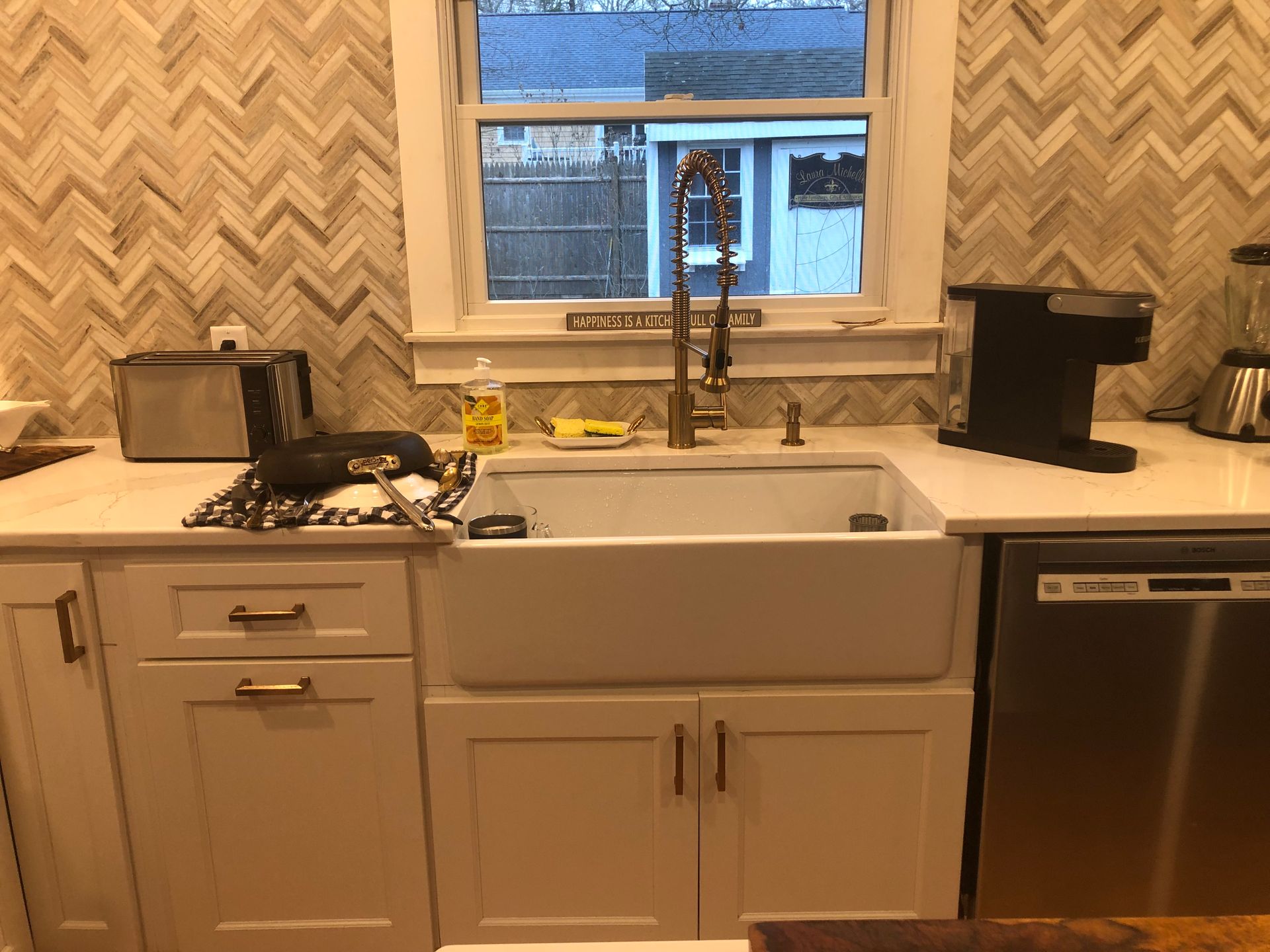 Kitchen with white cabinets, farmhouse sink, gold faucet, dishwasher, and herringbone backsplash.