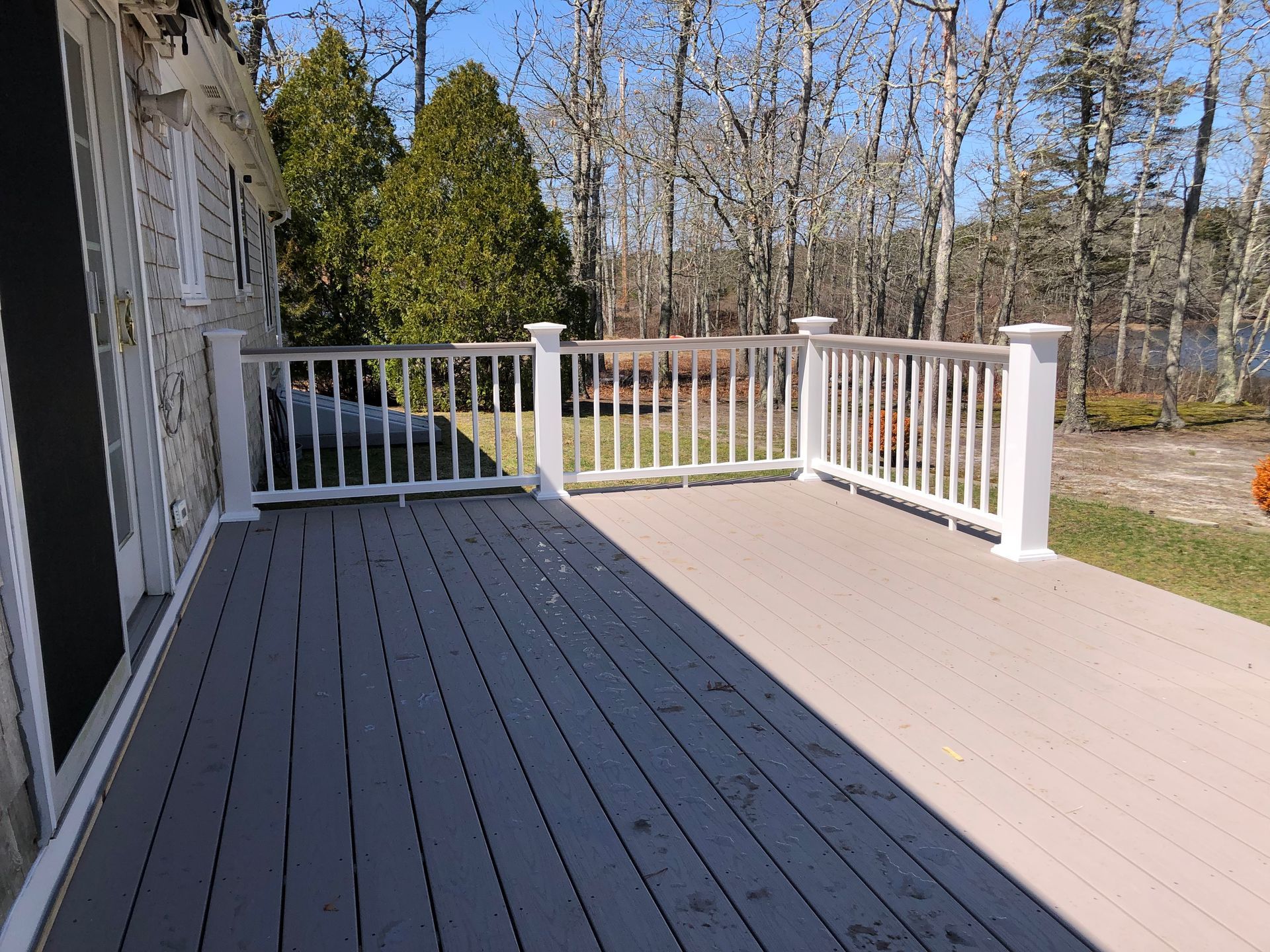 A wooden deck with white railing, overlooking a wooded area. The deck is bathed in sunlight.
