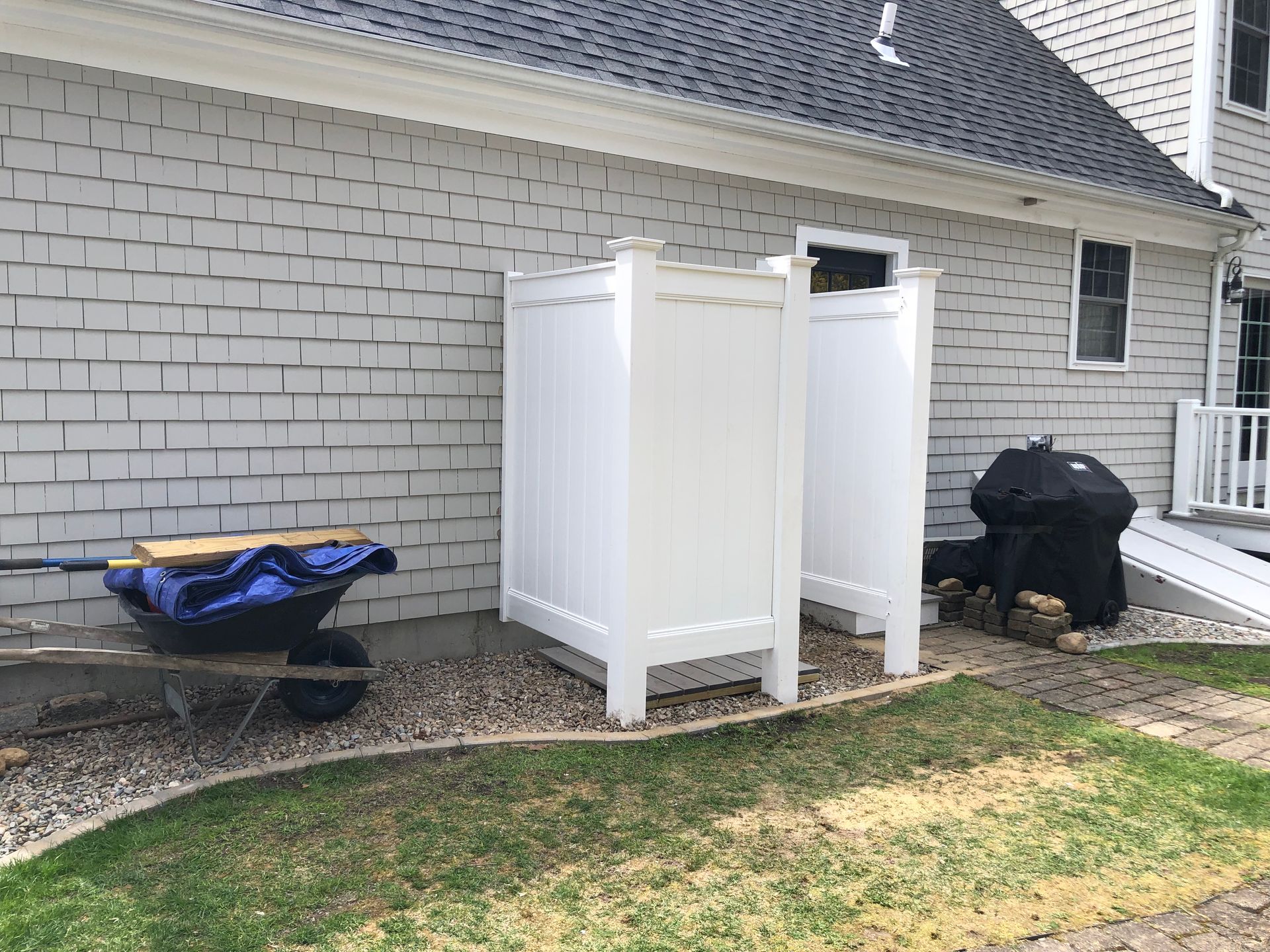 White outdoor shower structure next to a house with a grill and landscaping.
