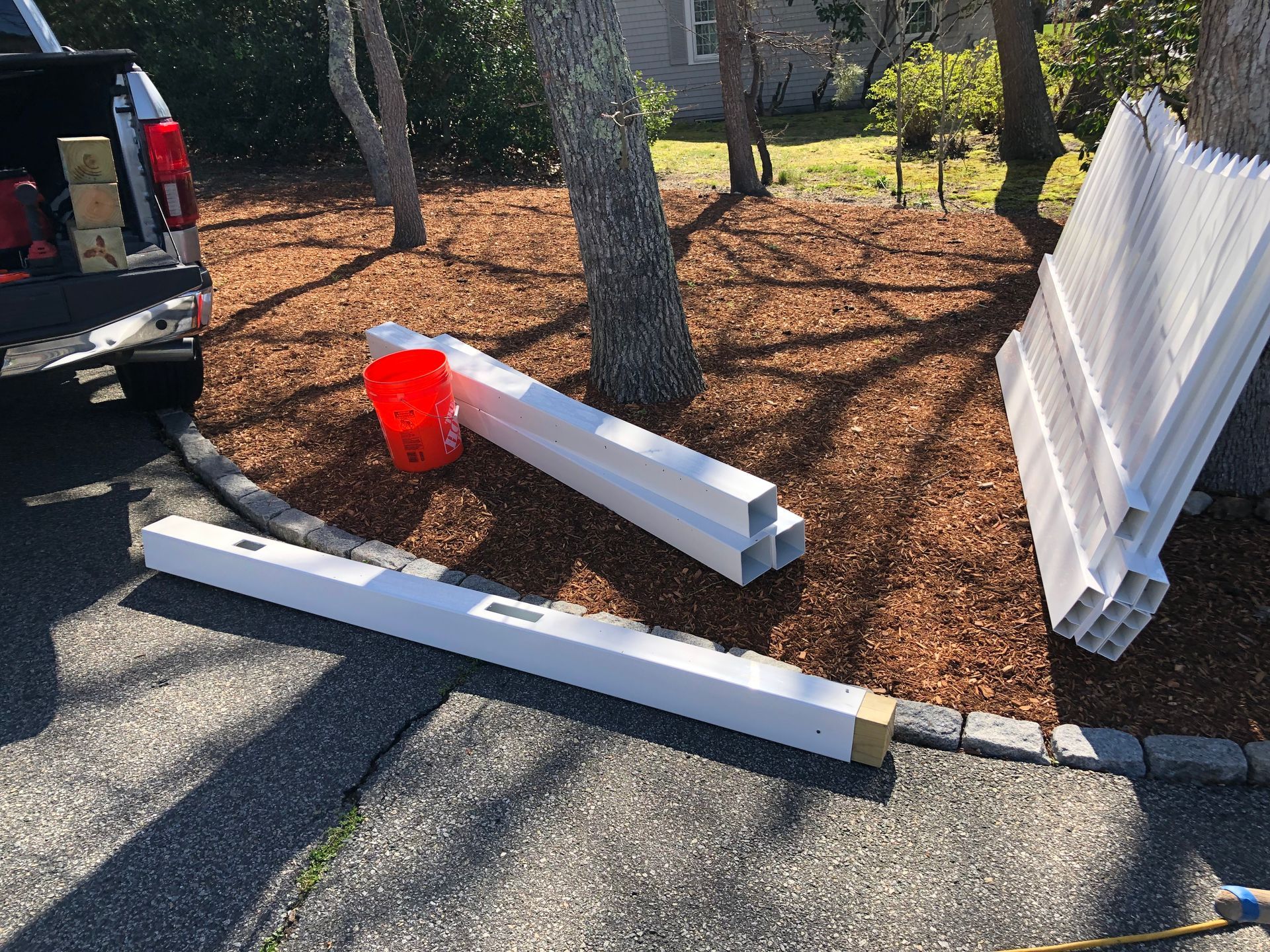 White fence materials and truck bed on a driveway, with a red bucket and trees.