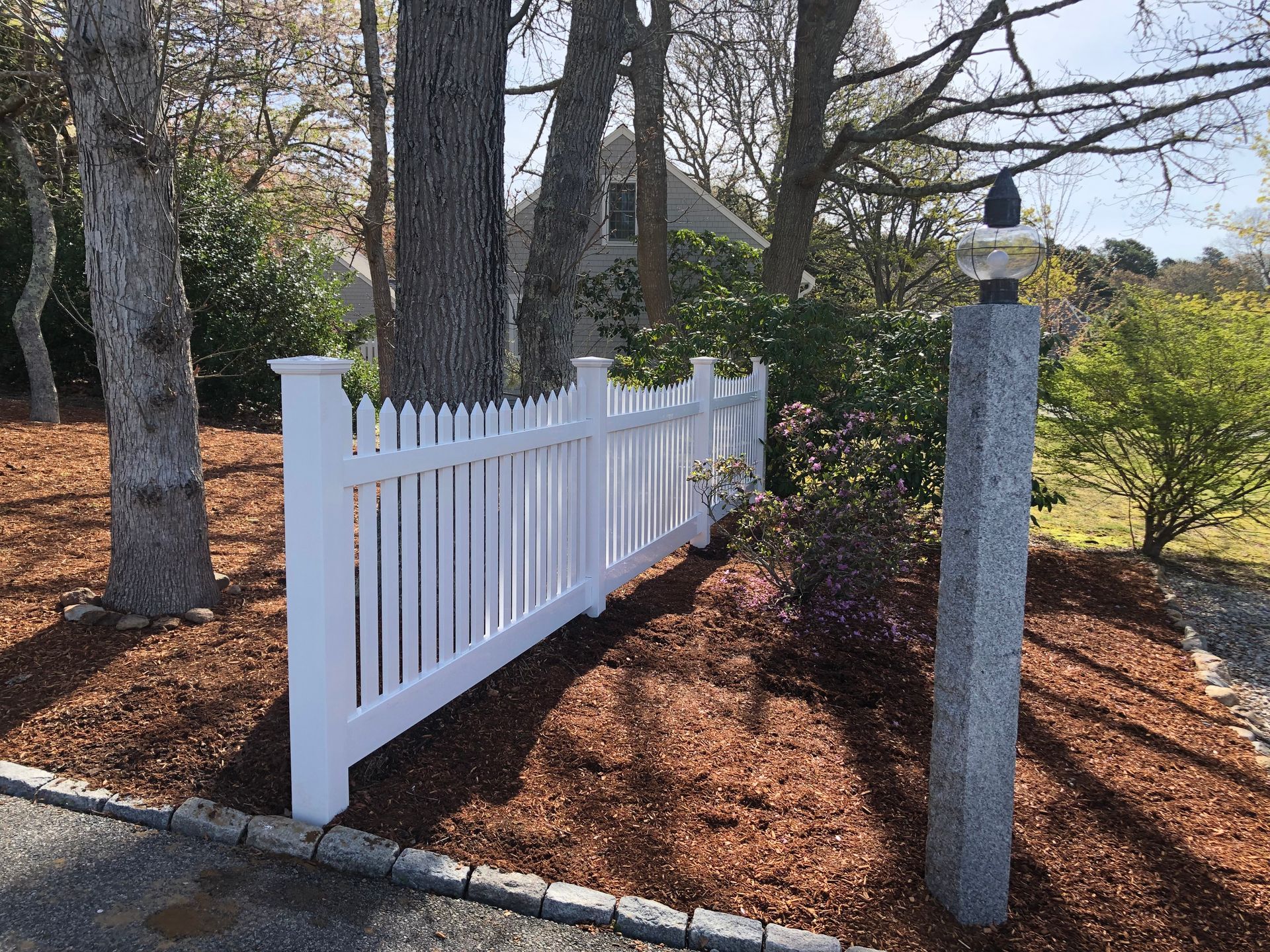 White picket fence with granite post lamp in front of a house, surrounded by trees and bushes.