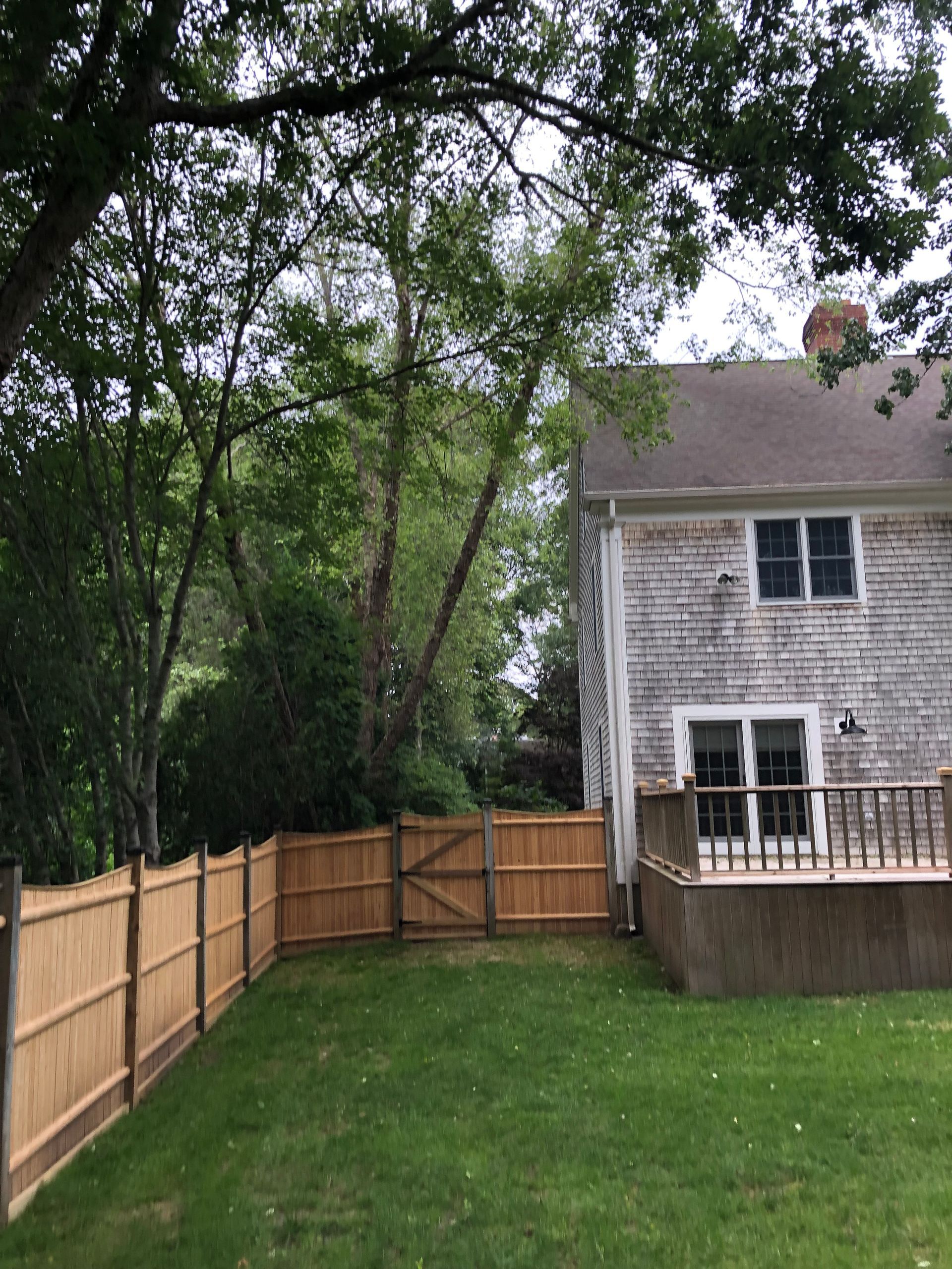 Wooden fence surrounds a grassy backyard, with a house on the right and trees in the background.