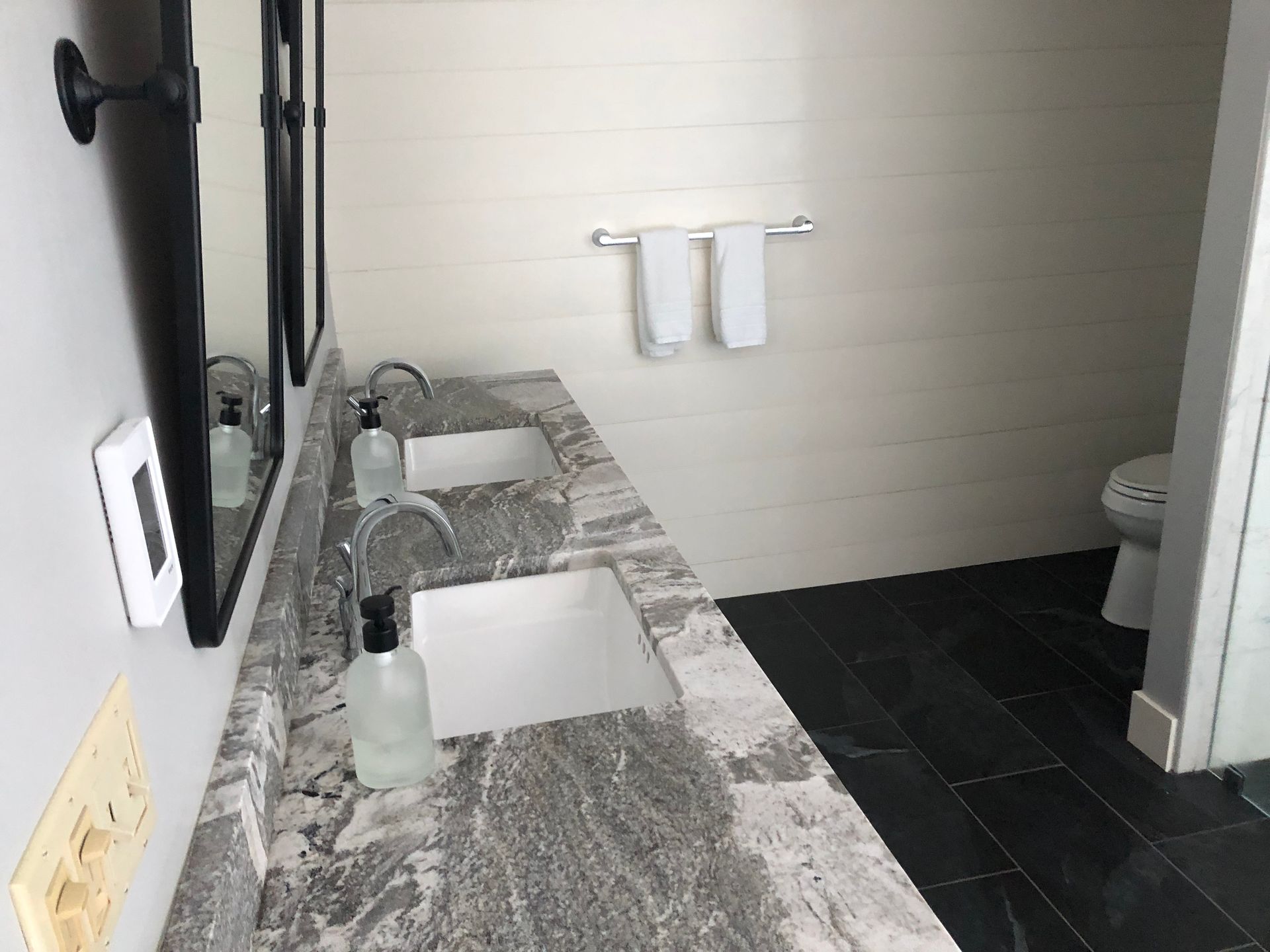 Bathroom with dual sinks, marble countertop, black floor tiles, white shiplap walls, and toilet.