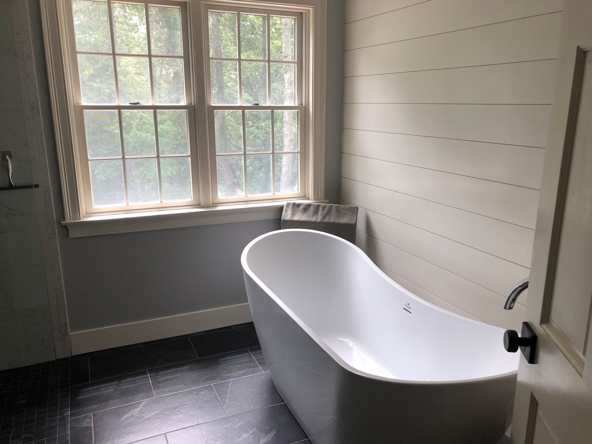 Freestanding white bathtub in a bathroom with window, gray walls, and black floor tiles.