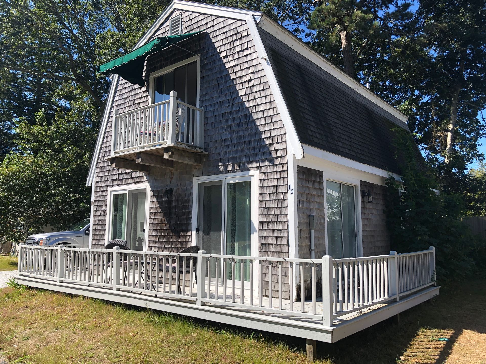 Cottage with gray cedar shingles, balcony, and deck, surrounded by trees and grass on a sunny day.