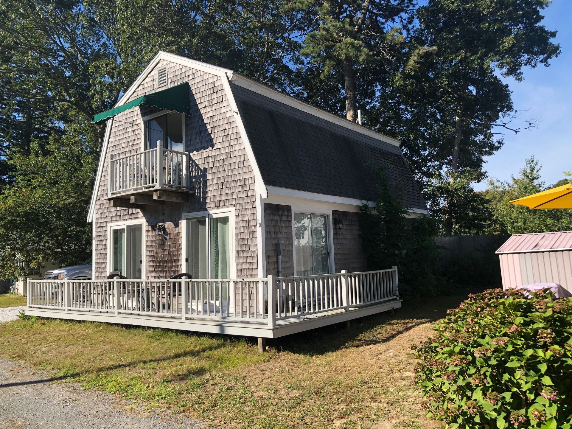 Cottage with a gray shingle roof, balcony, and porch. Green awning over an upper window. Surrounded by trees and grass.