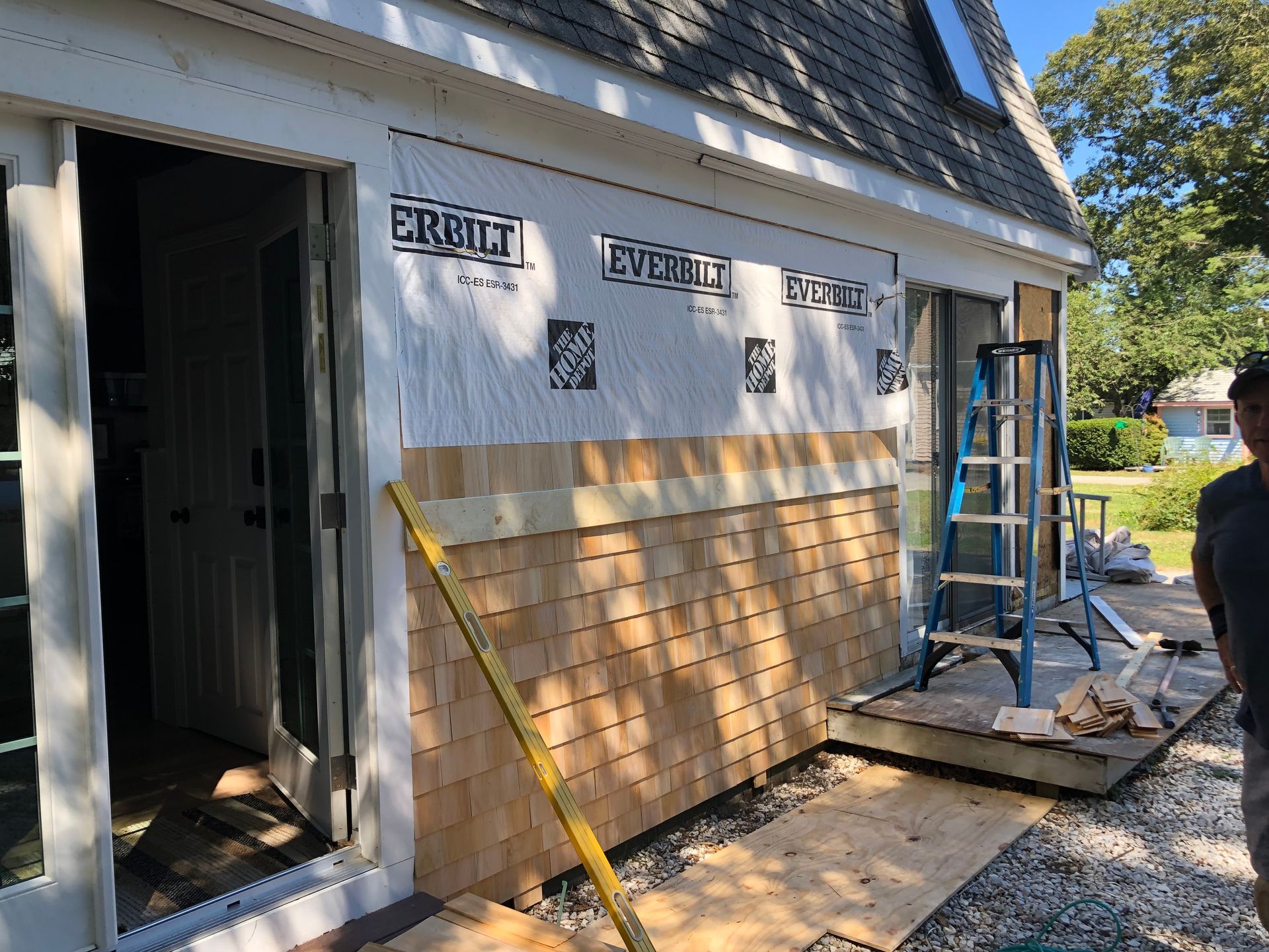 Construction site with cedar shingles being installed on a house, with a ladder and a person visible.