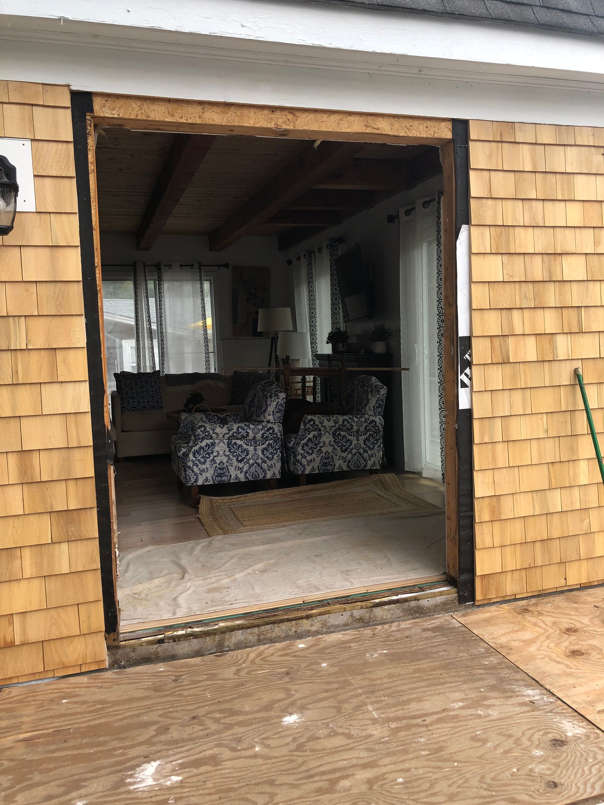 Exterior view of a house opening. Brown shingles surround an open doorway leading to a living room with patterned chairs.
