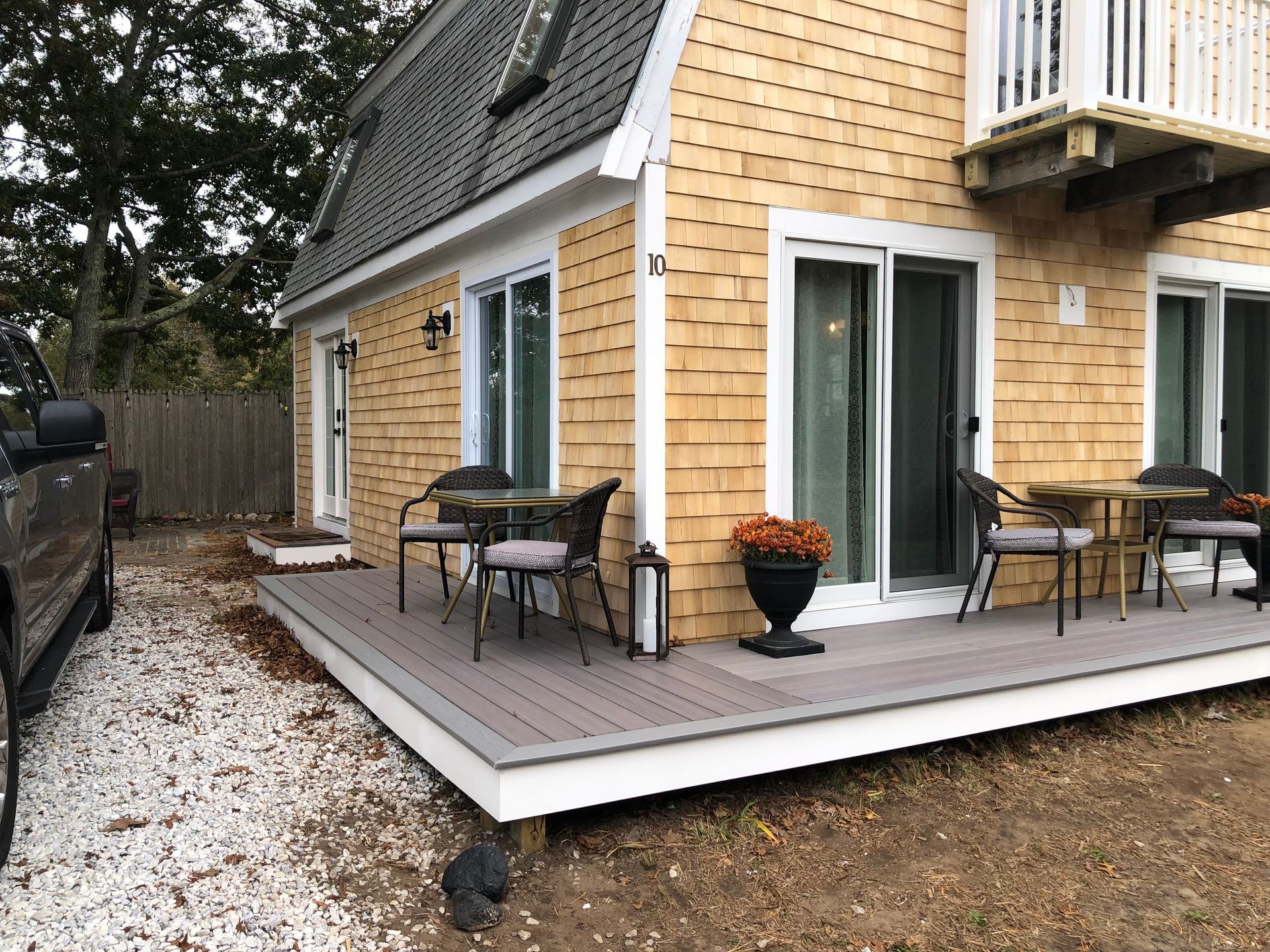 Cottage with a gray deck, two sliding doors, and light brown shingled exterior. Two small tables with chairs on the deck.