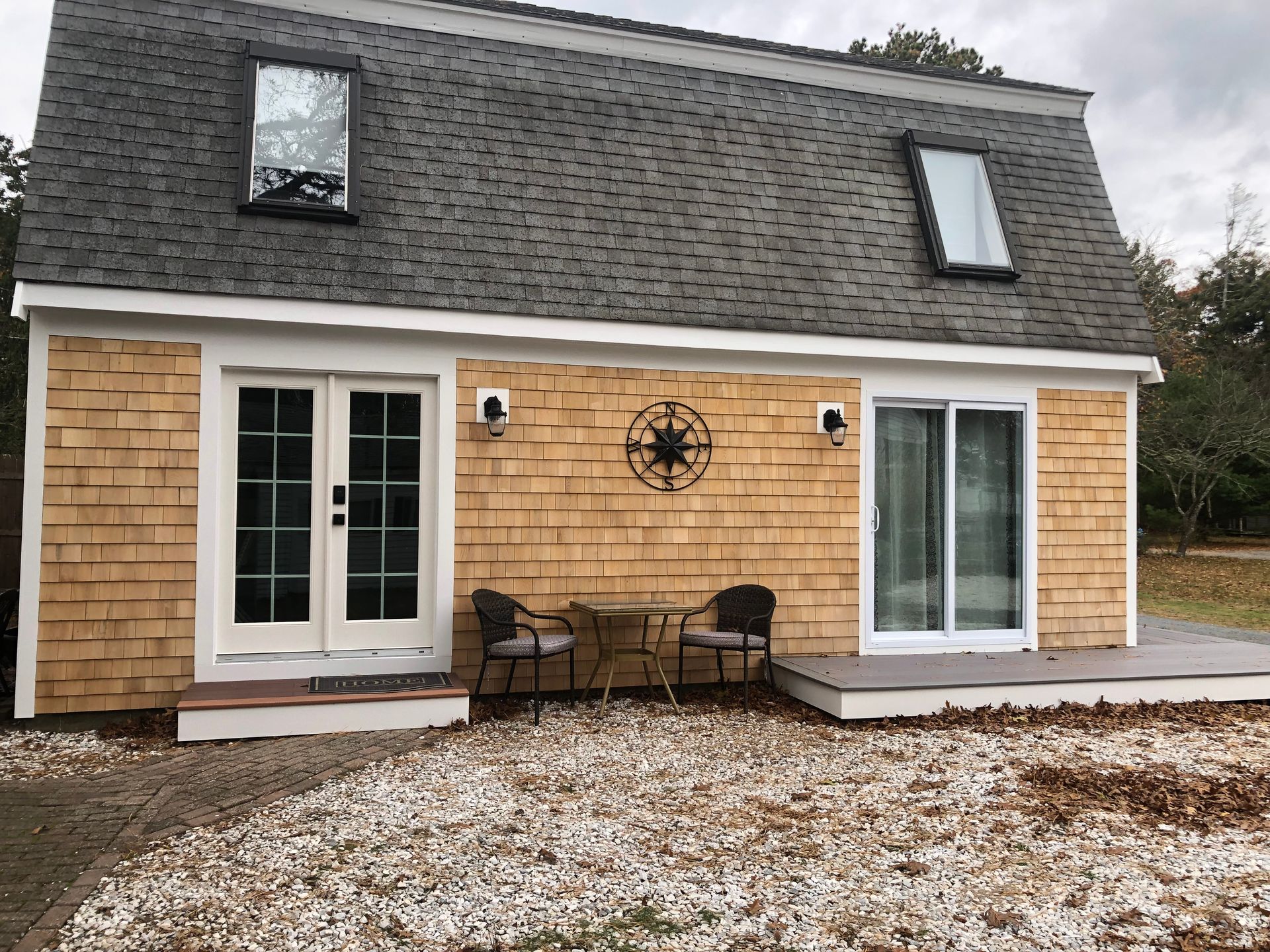 Small cottage with shingle siding, two dormers, French doors, and a sliding glass door. Two chairs sit outside.