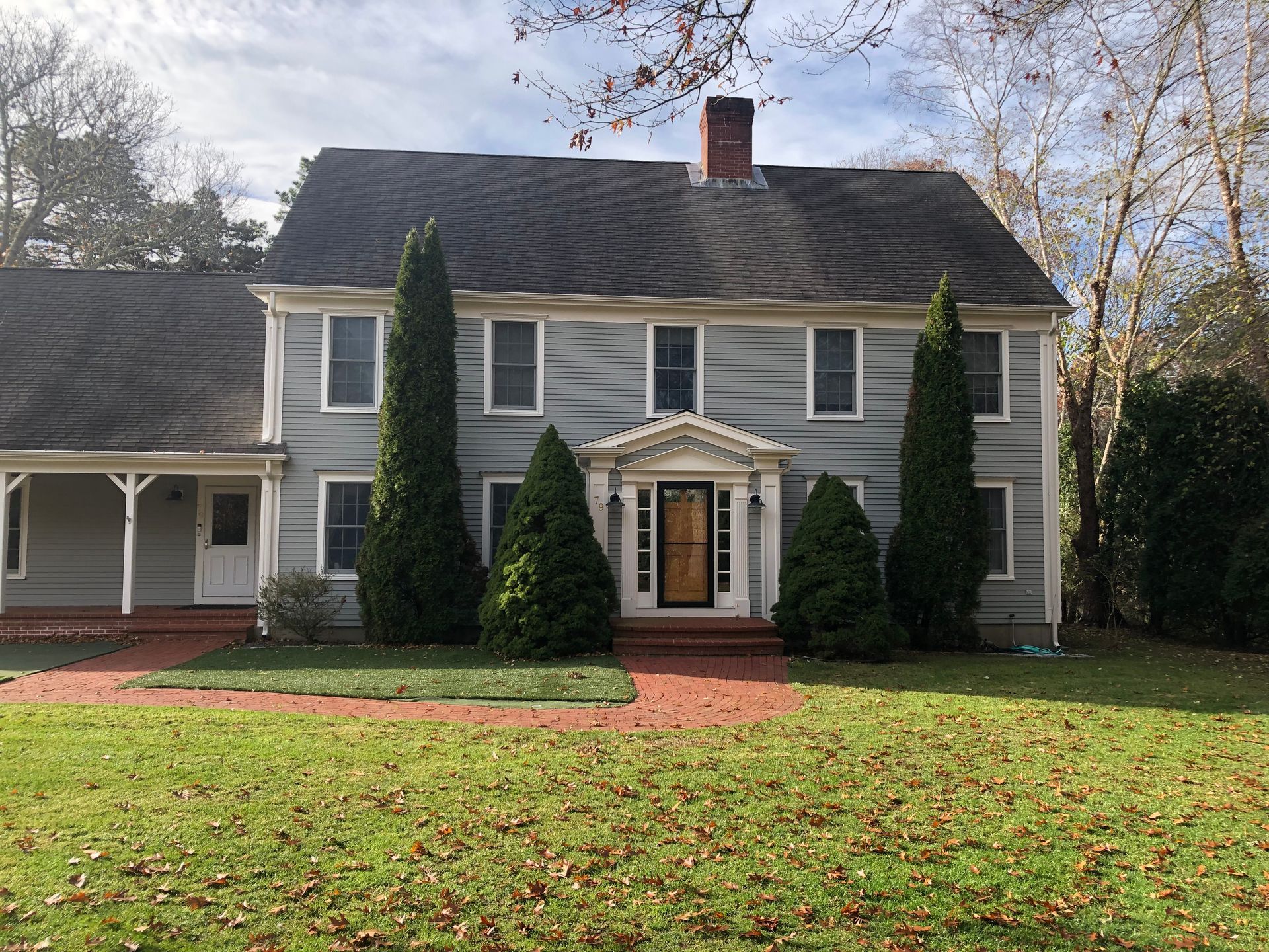 Gray two-story house with black roof, brick walkway, and green lawn. Tall trees flank the front door.
