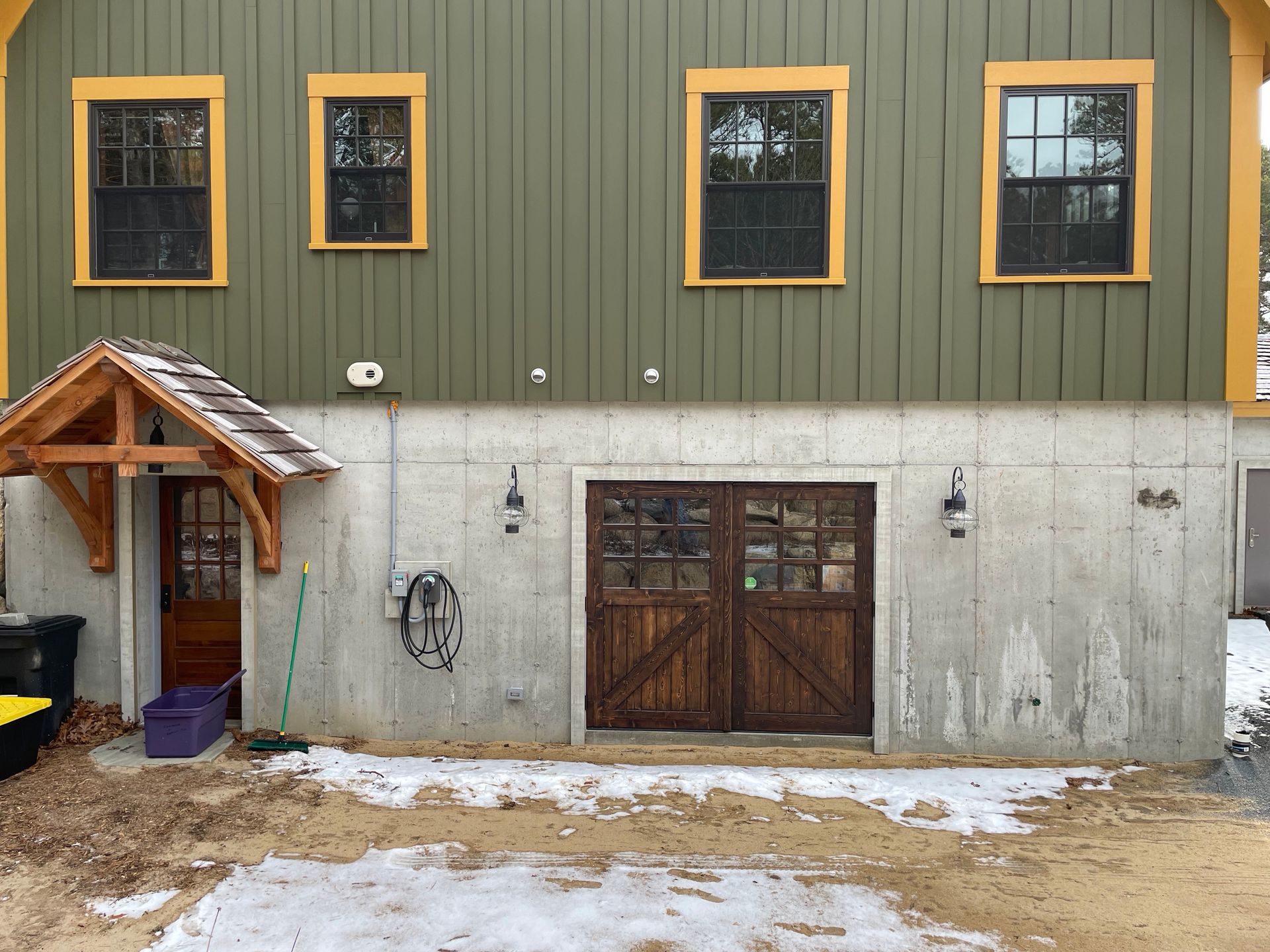 Green building with wooden doors and windows, concrete foundation. Snow and dirt on the ground.