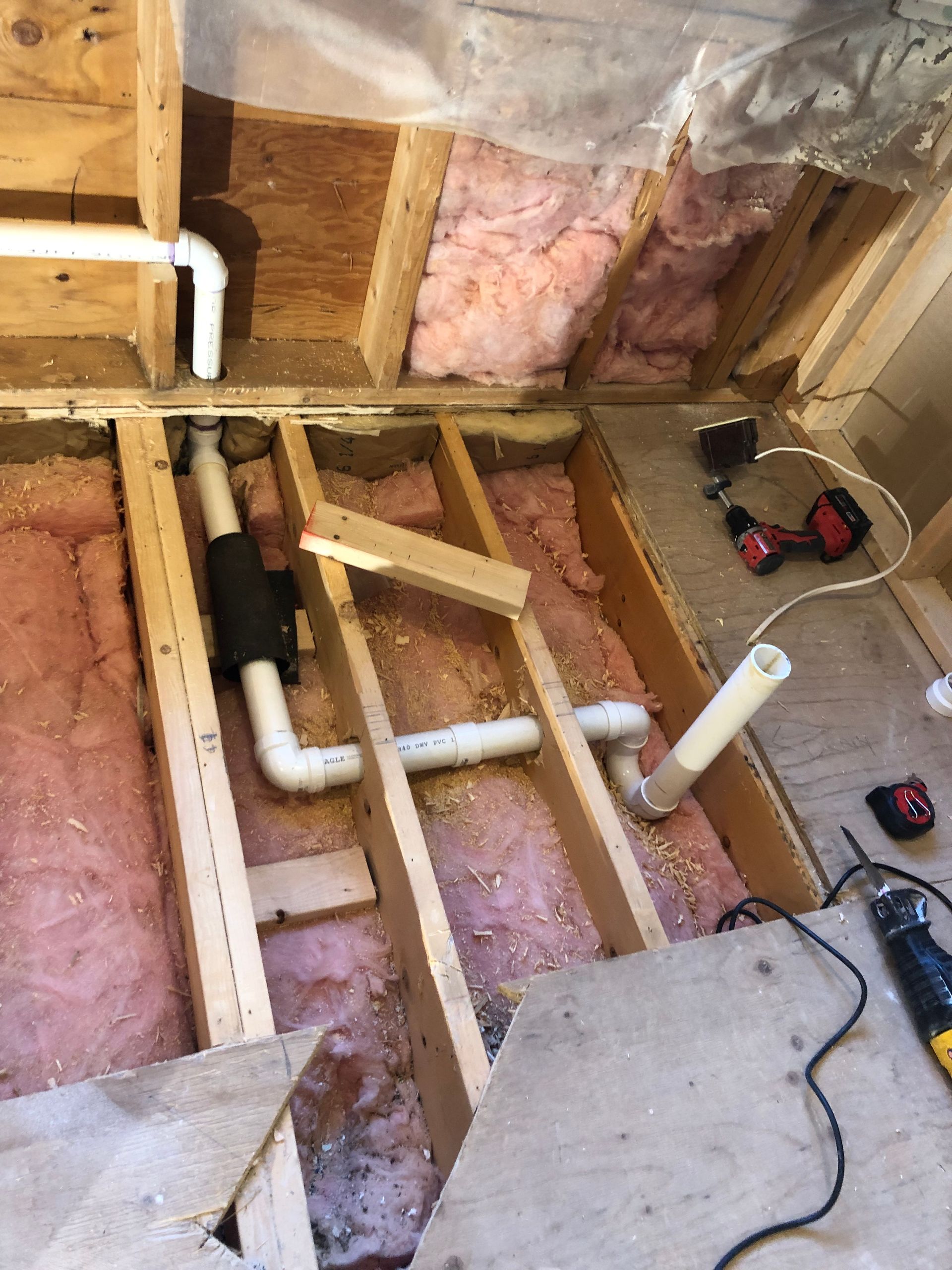 Plumbing pipes installed in a wooden floor frame, with insulation visible. A drill and saw are nearby.