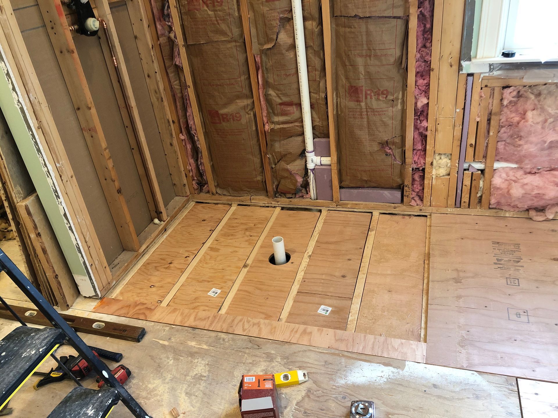 Bathroom under construction: exposed studs, plywood floor with drain, insulation visible.