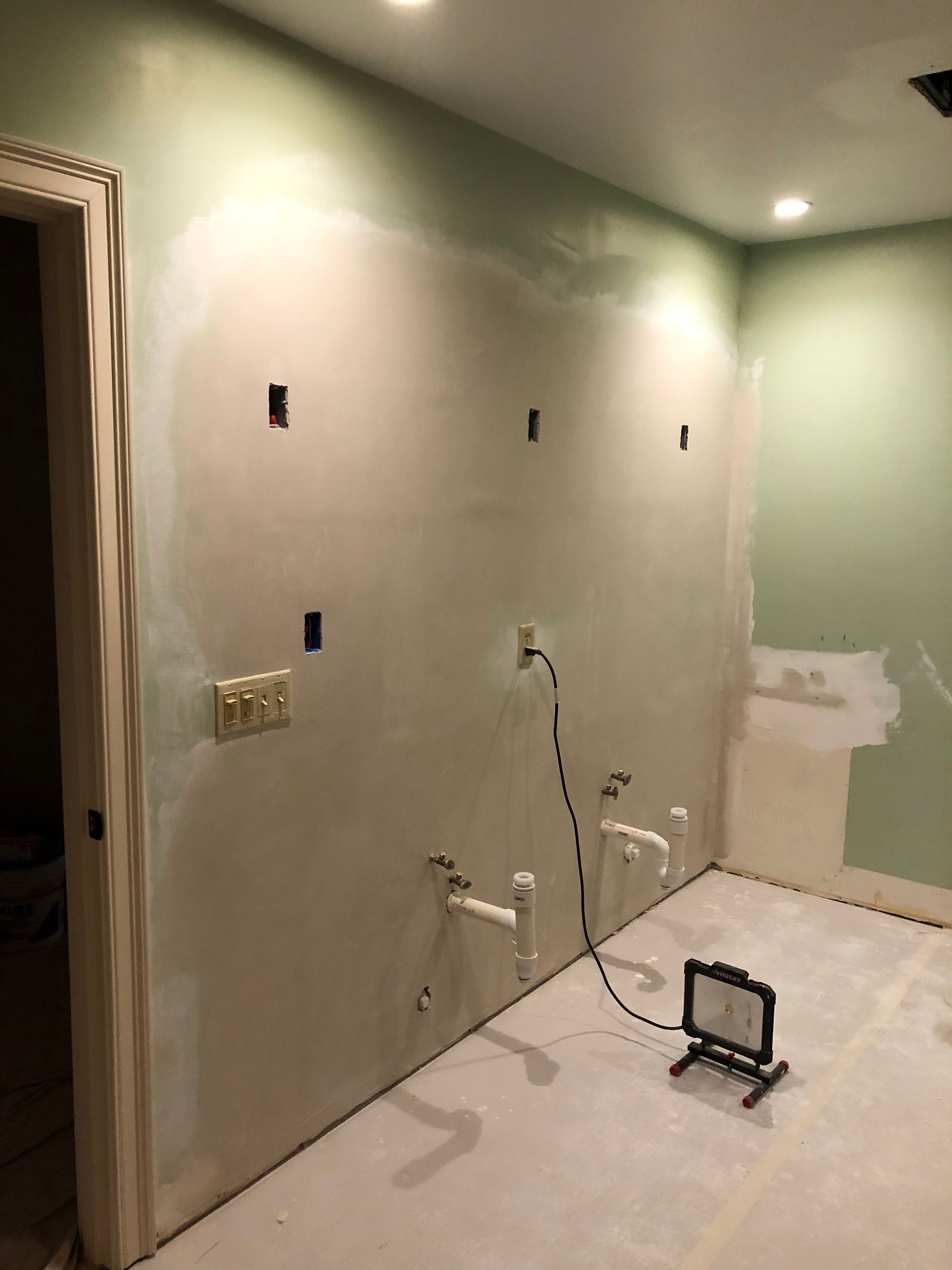 Bathroom under construction; walls are plastered and painted green, with exposed plumbing and electrical outlets.