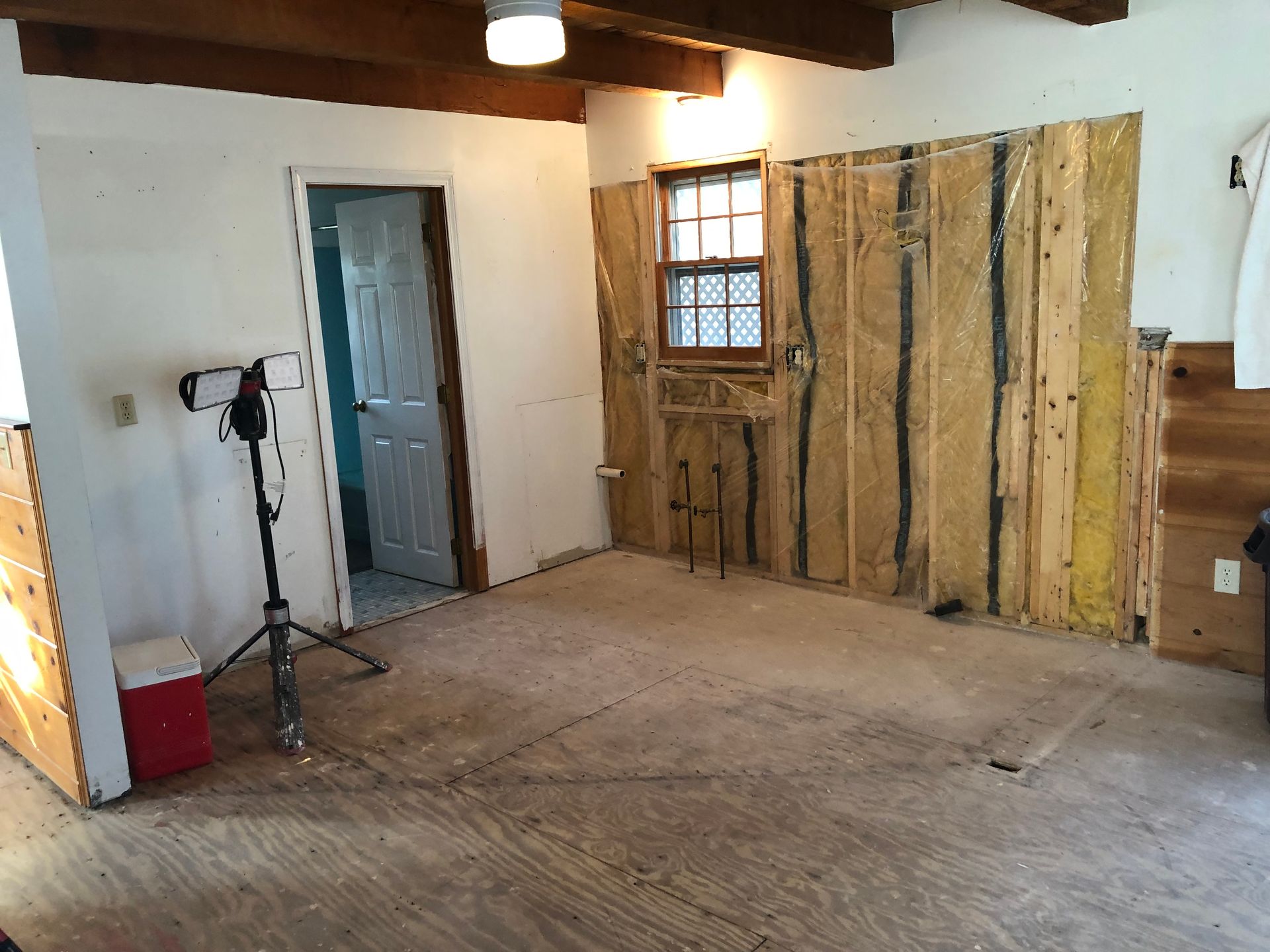Interior room renovation in progress: exposed wall studs, window, open doorway, plywood floor.