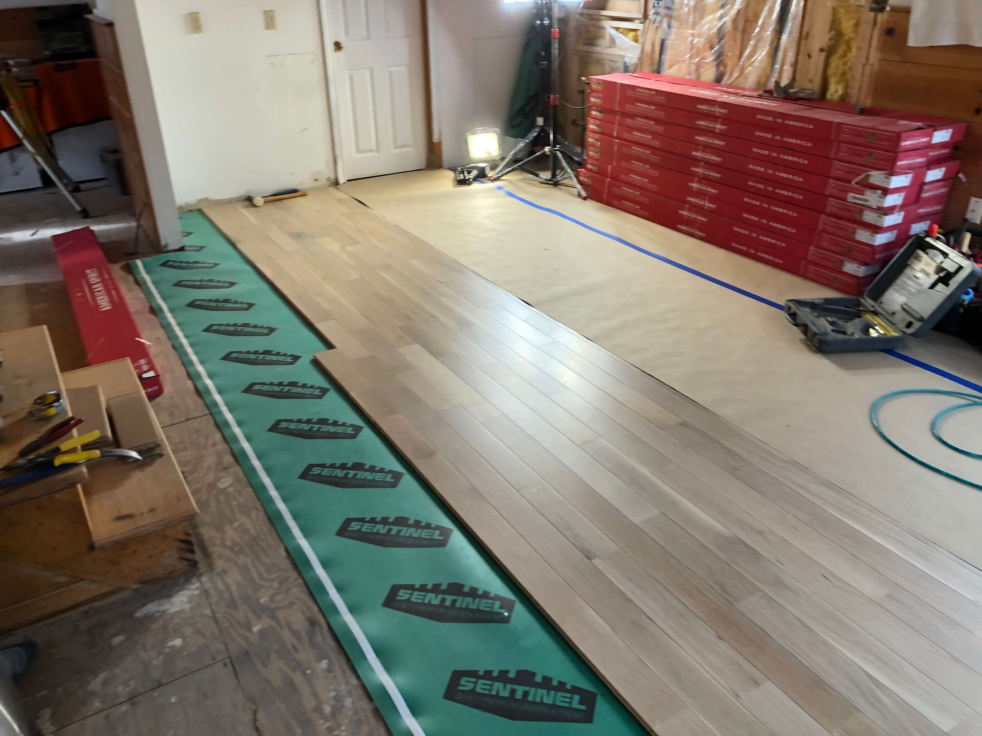 Hardwood flooring installation in progress; wood planks laid on green underlayment, tools, and stacked boxes.