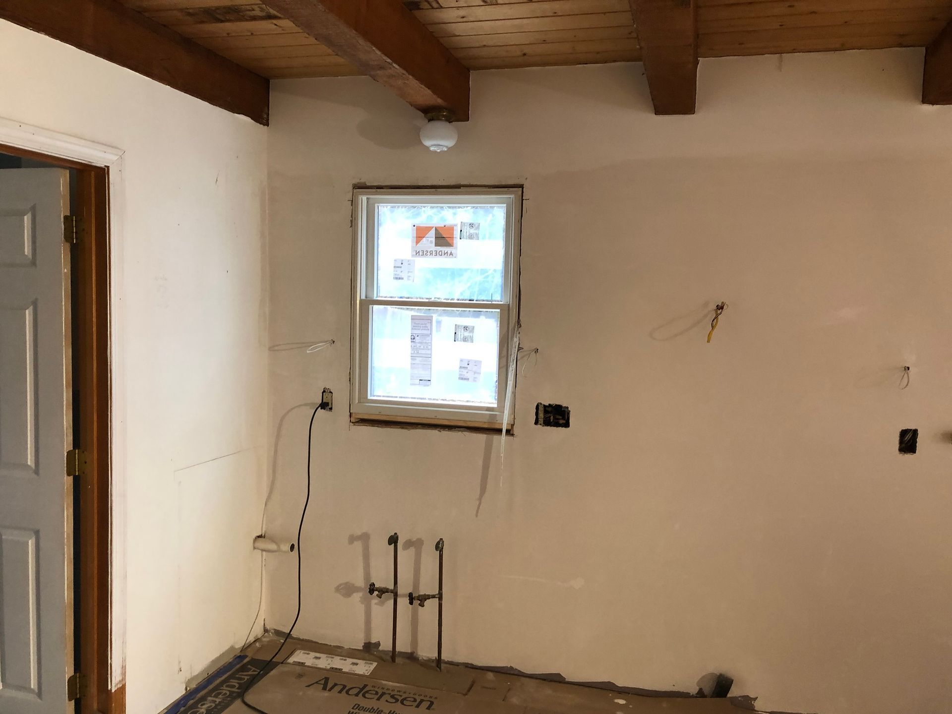 Interior of a room under renovation: unfinished walls, window, exposed wiring, wooden beams on the ceiling, and a door.