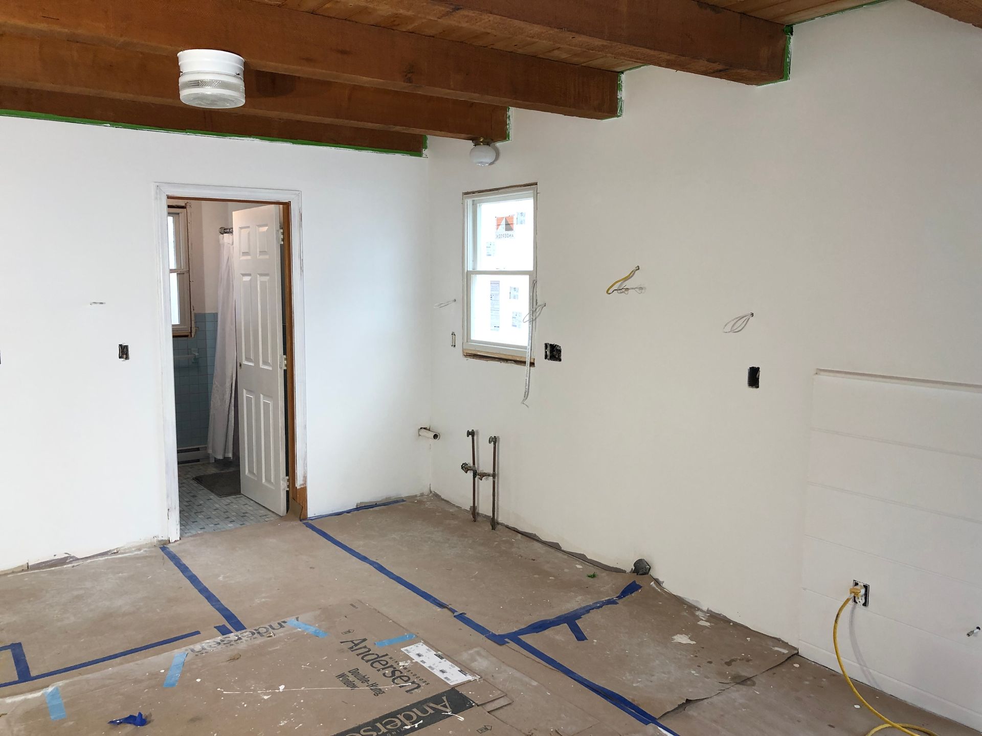 Interior of a room under renovation: white walls, exposed wooden beams, open doorway, window. Blue tape outlines on floor.