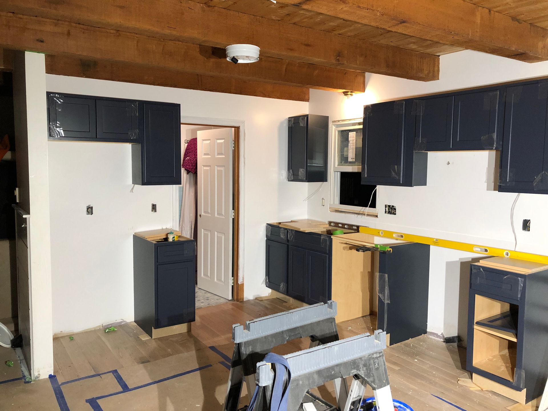 Kitchen renovation in progress, dark blue cabinets installed on white walls, exposed wooden beams on ceiling.