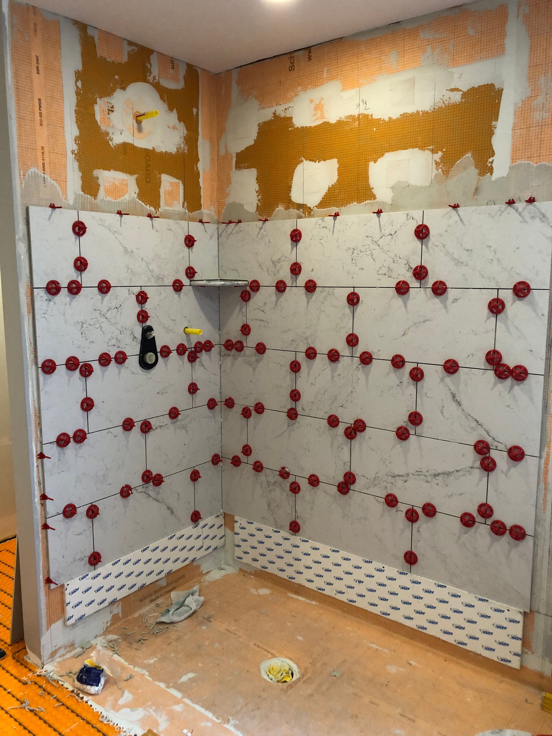 Shower walls in progress with large marble-like tiles and red leveling clips, a corner shelf.