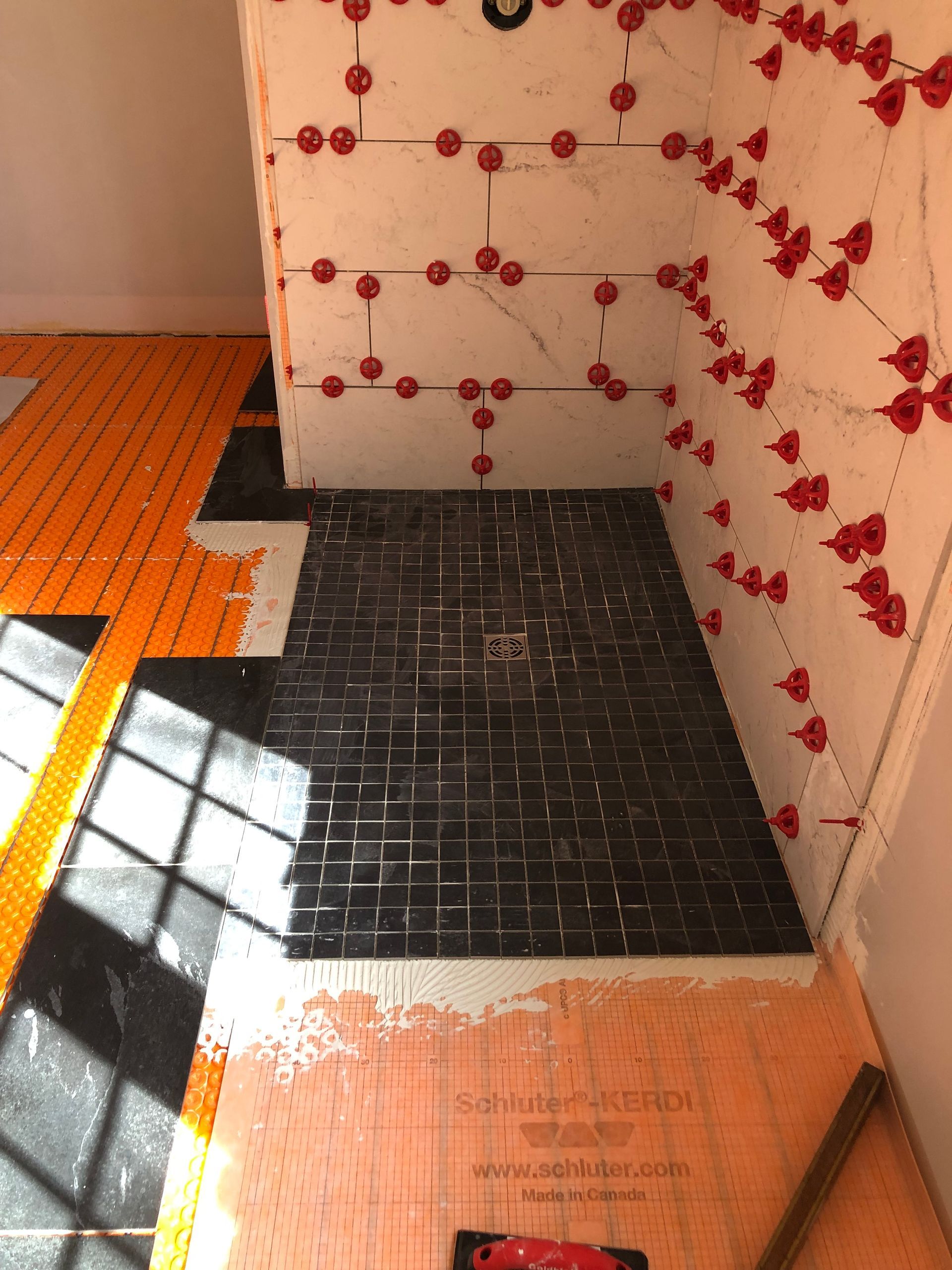 Bathroom under construction, showing shower floor with dark tiles and surrounding walls. Orange heated flooring is visible.