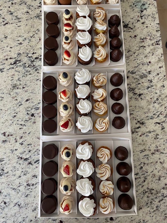 Pasteles variados en tres filas, incluyendo galletas, mini pasteles y bolas de chocolate, en una caja blanca sobre una mesa.