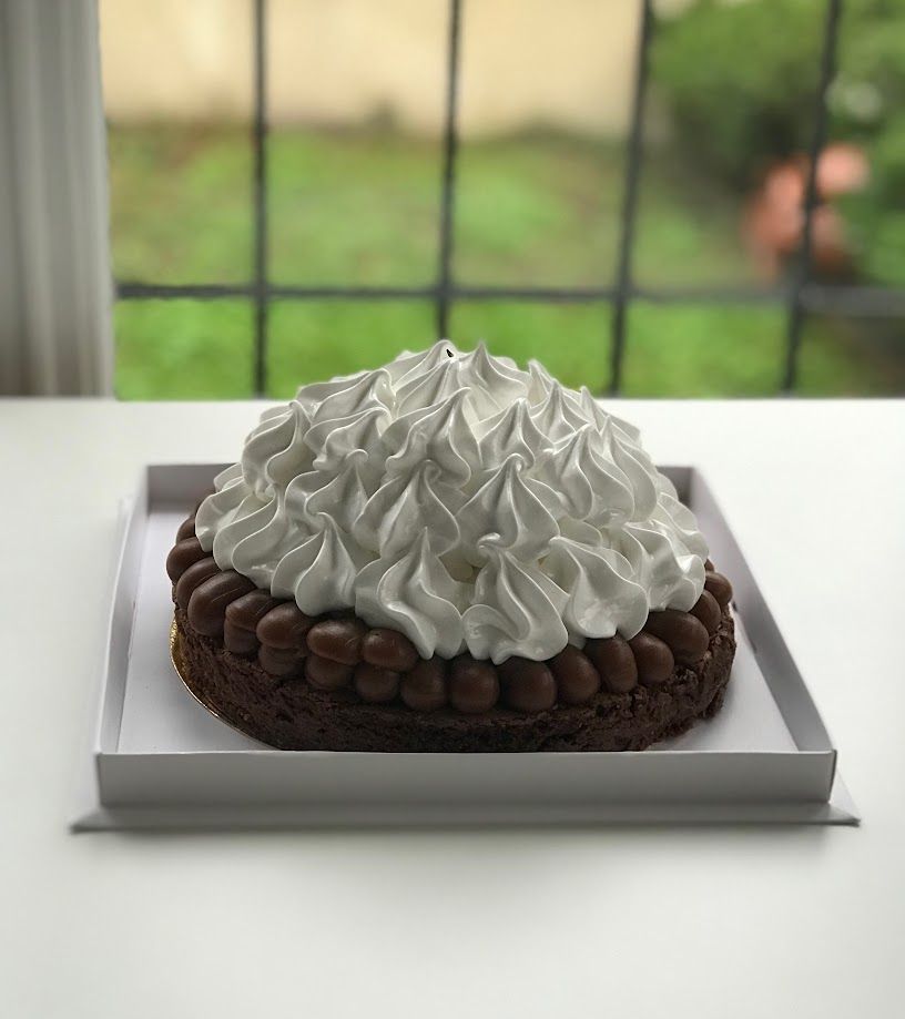 Pastel de chocolate con glaseado de merengue, presentado en una caja blanca sobre una mesa blanca. Ventana al fondo.
