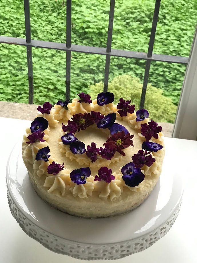 Pastel decorado con flores de color violeta y glaseado color crema, sobre un soporte para pastel blanco, frente a una ventana.