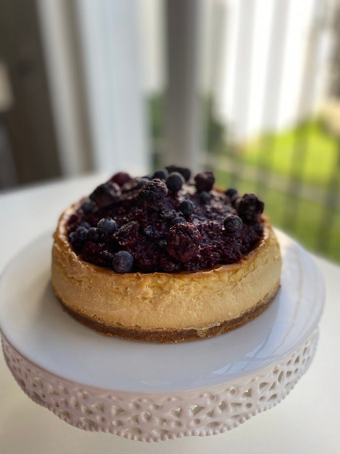 Tarta de queso con cobertura de frutos rojos sobre un soporte para tartas blanco, junto a una ventana luminosa.