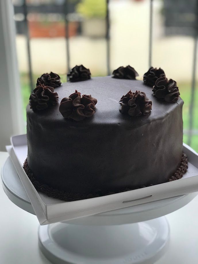Pastel de chocolate con glaseado de chocolate y rosetas sobre un soporte para pastel blanco.