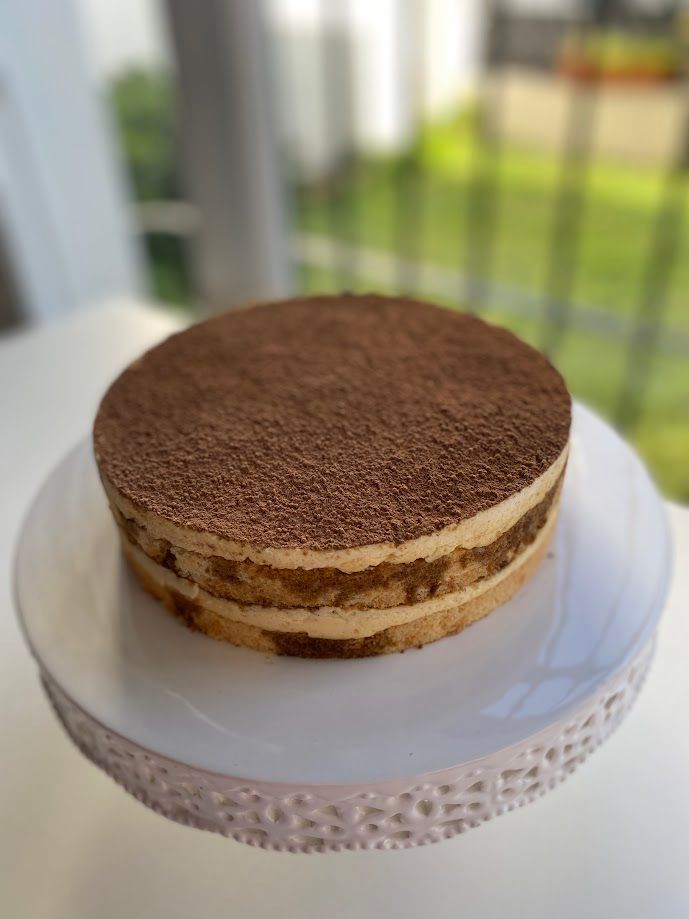 Pastel de tiramisú sobre pedestal blanco, con capas visibles y espolvoreado con cacao en la parte superior.