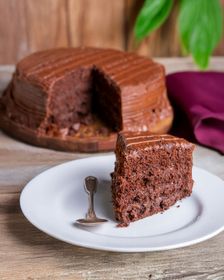 Rebanada de pastel de chocolate en un plato blanco, con el pastel entero al fondo. Mesa de madera.