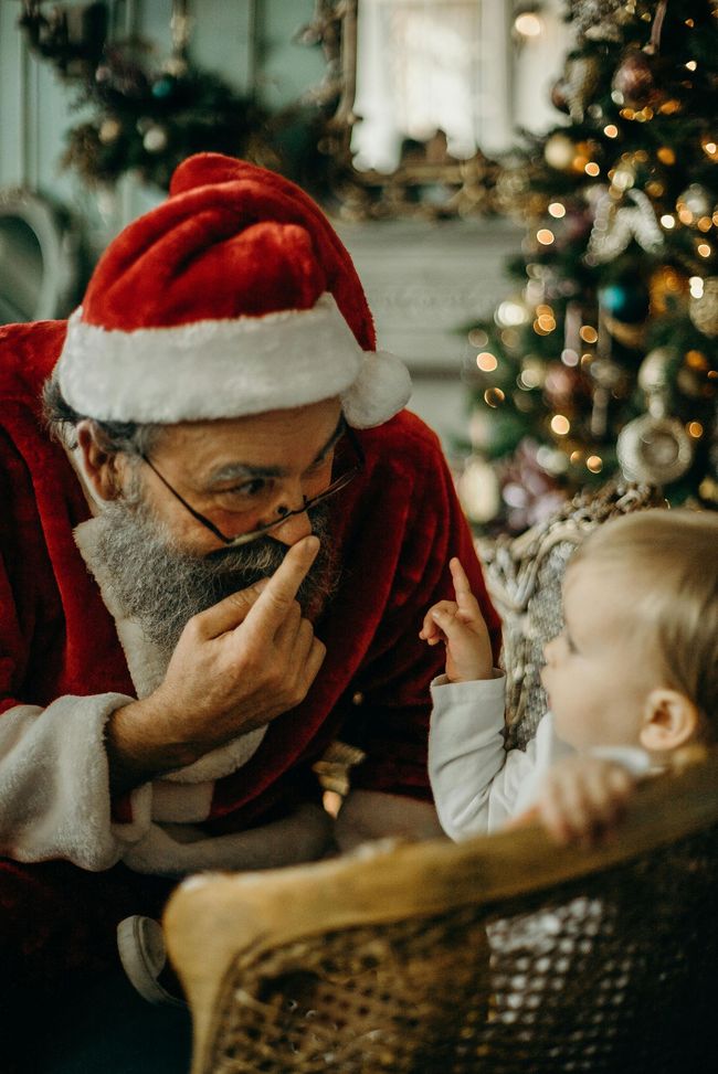 Santa Claus and a small child, indoors near a decorated Christmas tree, pointing fingers at each other.