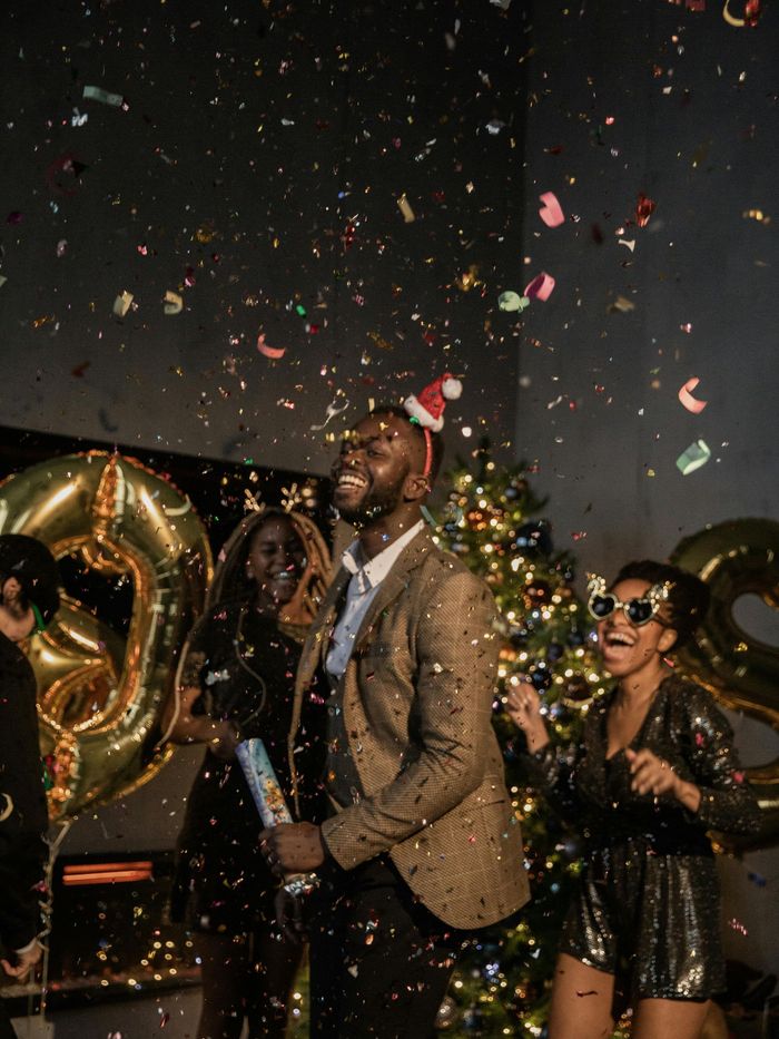 People celebrating, confetti falling. Man in blazer and Santa hat, others in festive attire, with gold balloons.
